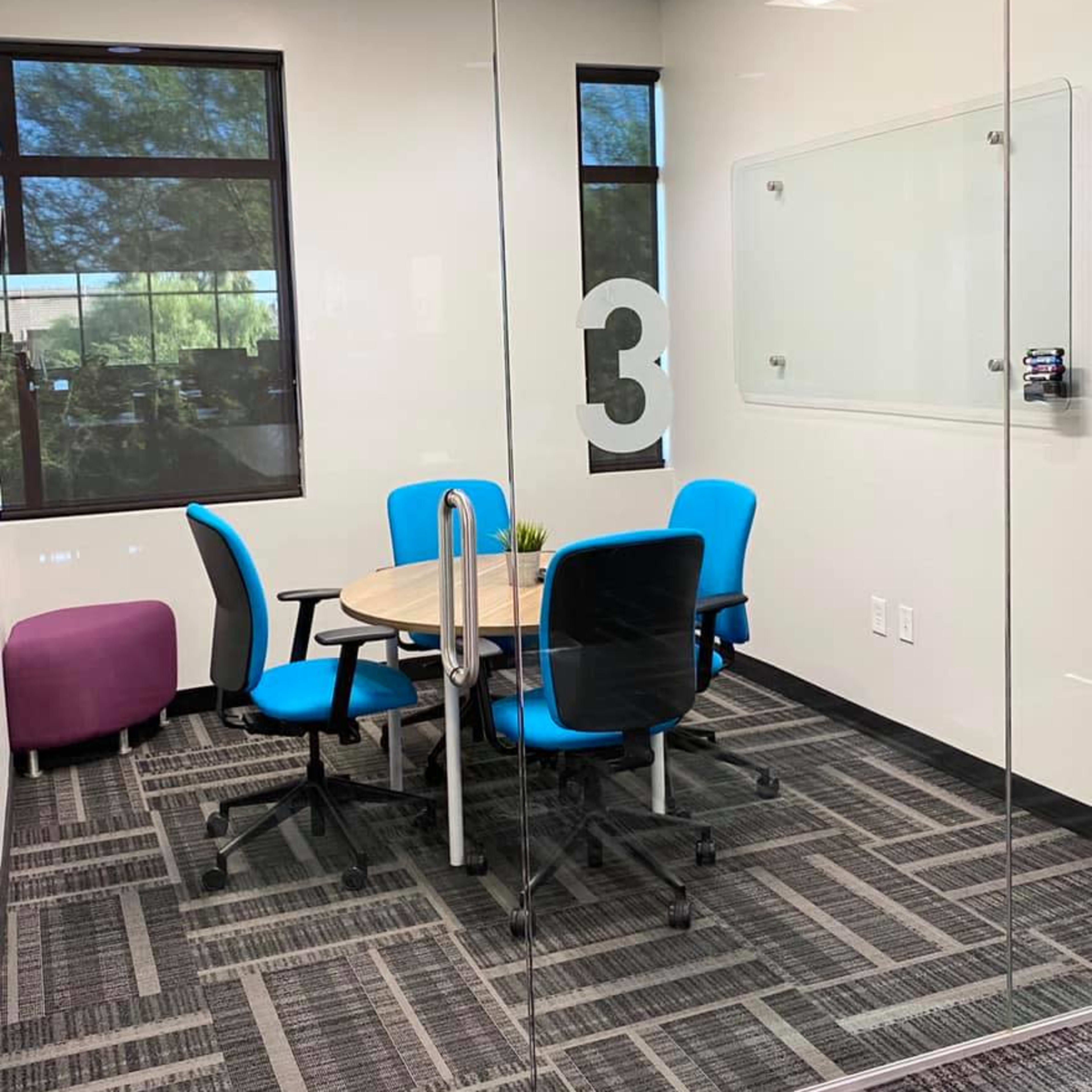 A small conference room features a round table surrounded by four blue chairs and a purple ottoman, with large windows allowing natural light.