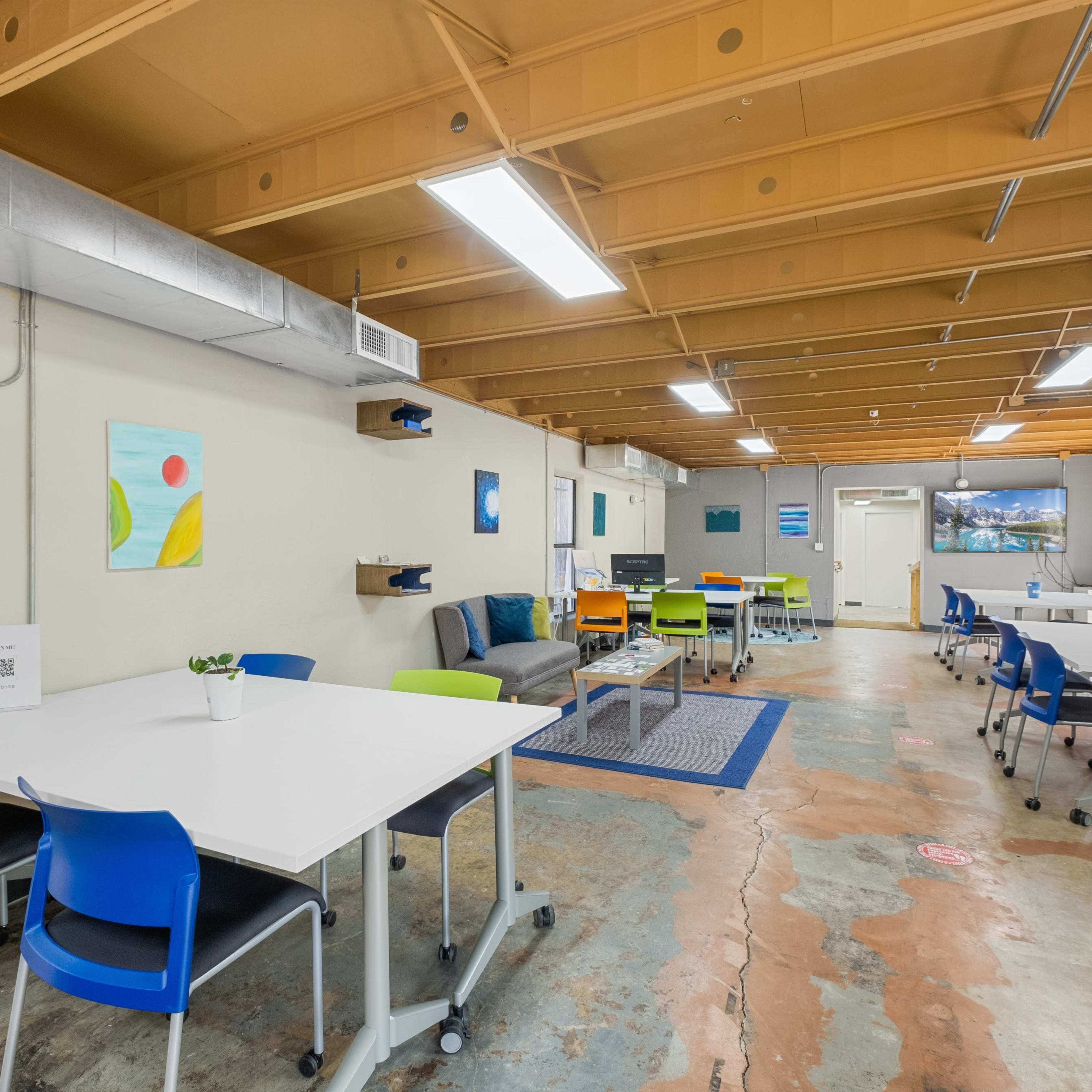 A spacious, well-lit office area with multiple tables and chairs, a television screen, and colorful wall art.