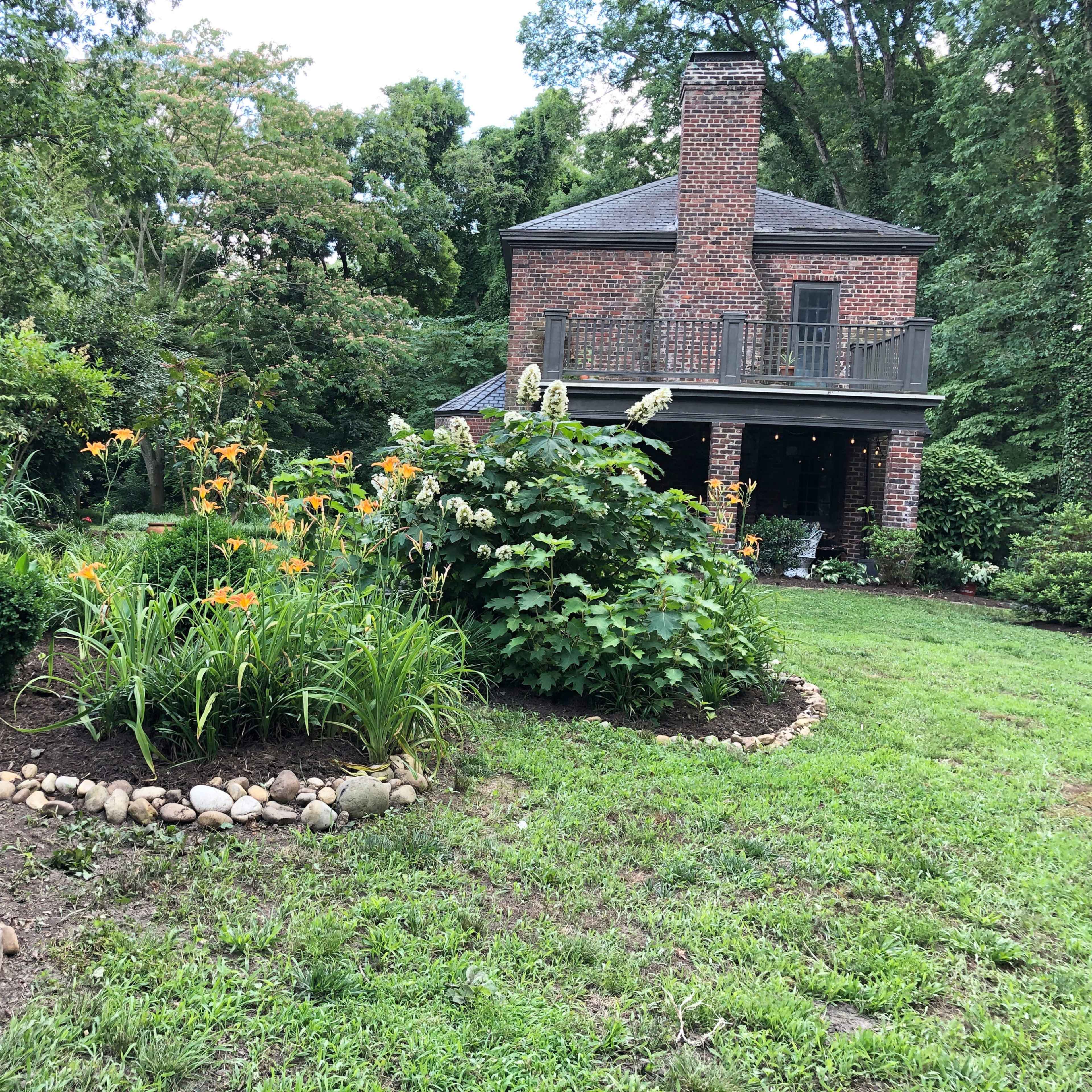 A two-story brick house is surrounded by a landscaped garden featuring flowers and a rocky border.