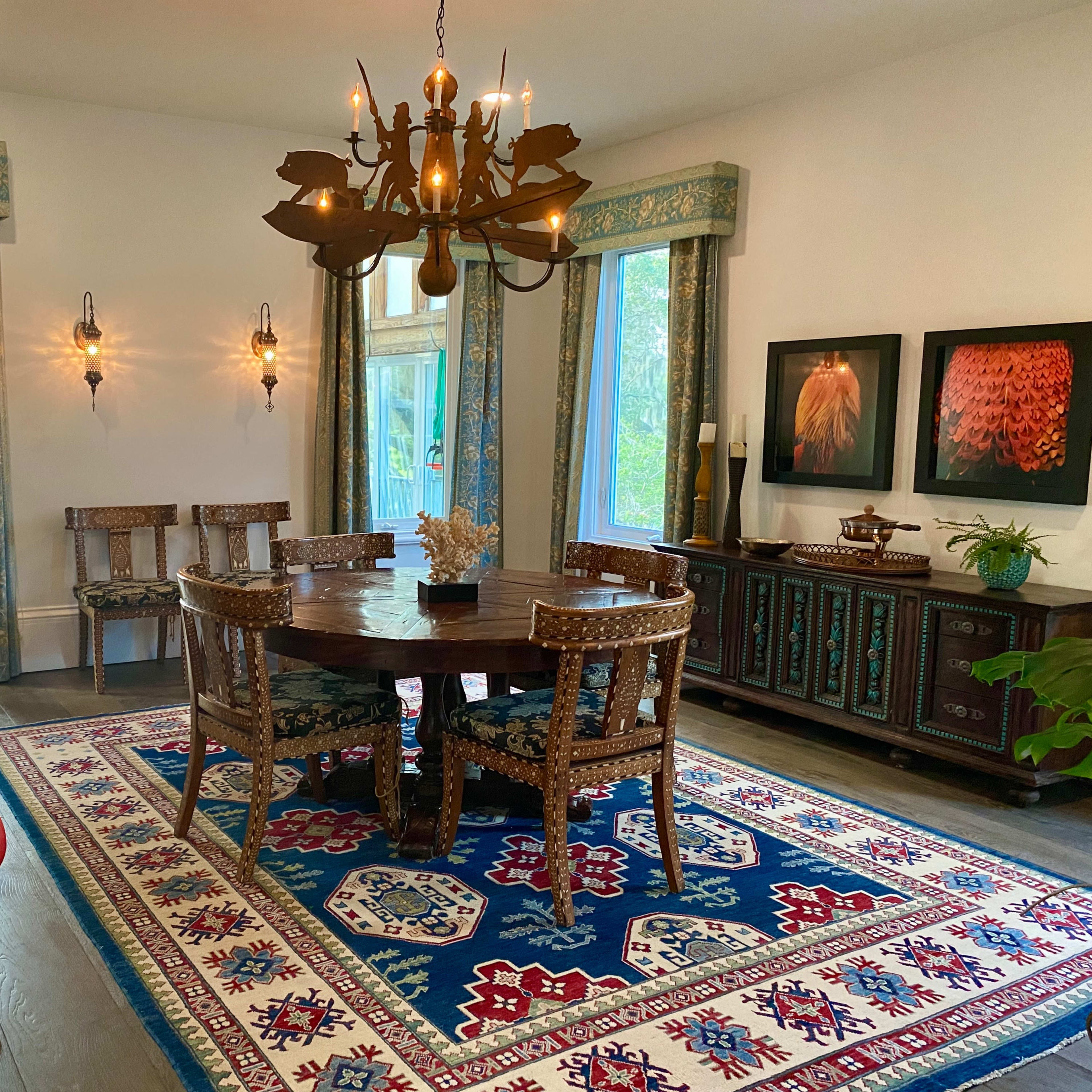 A dining area features a round wooden table surrounded by patterned chairs, illuminated by a chandelier, with a decorative rug and framed artwork on the walls.