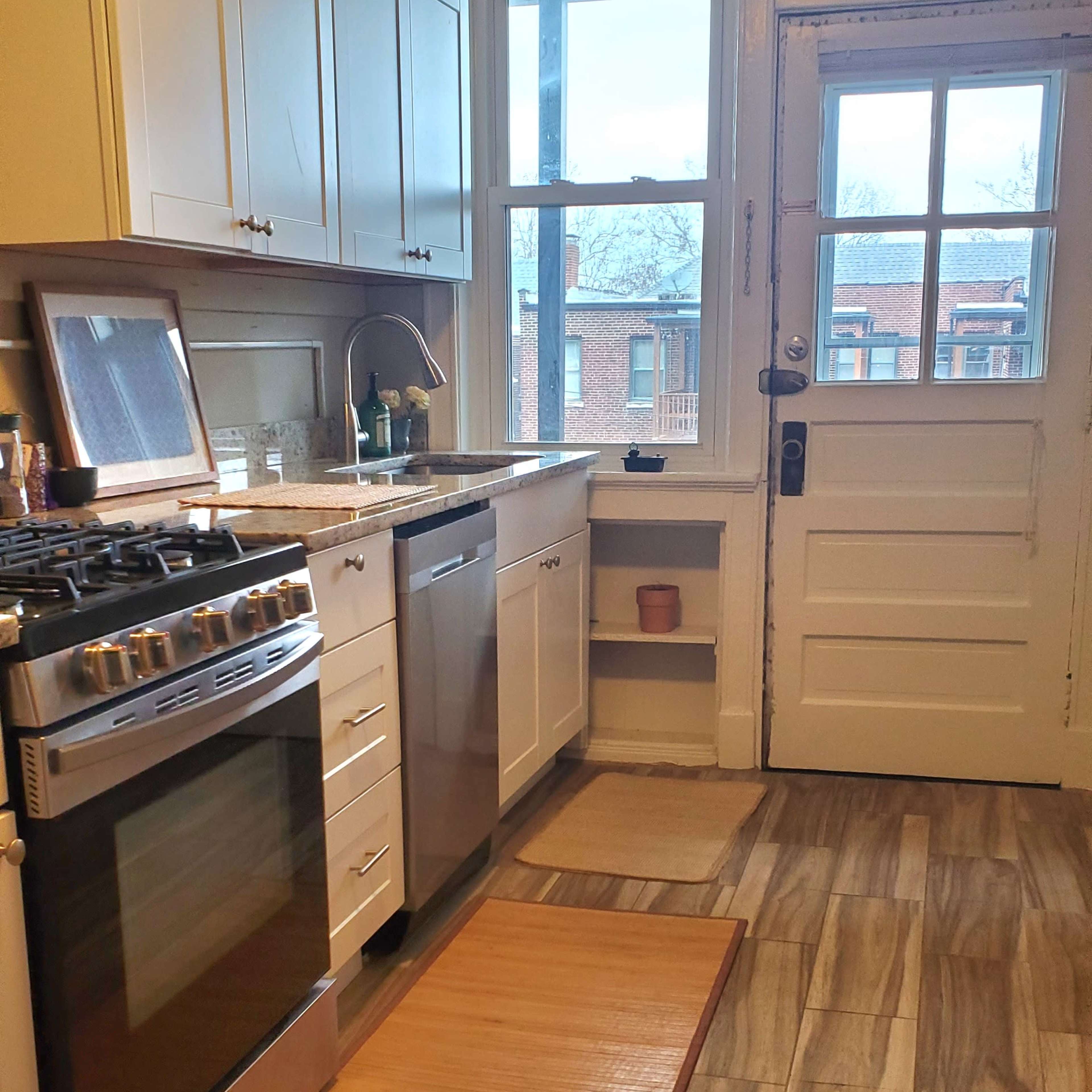 A kitchen features stainless steel appliances, white cabinets, and a door leading outside, with a wooden mat on the tiled floor.