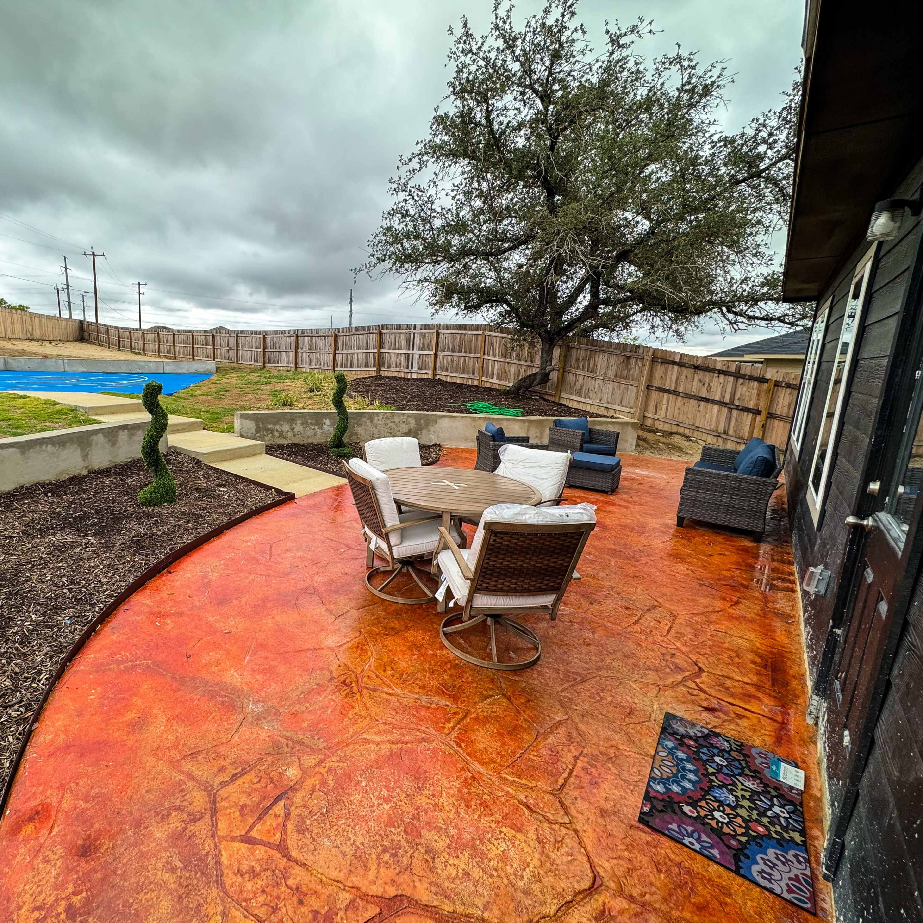 A backyard features a circular seating area with a table and chairs, surrounded by decorative shrubs, a basketball court, and a tree in the background.