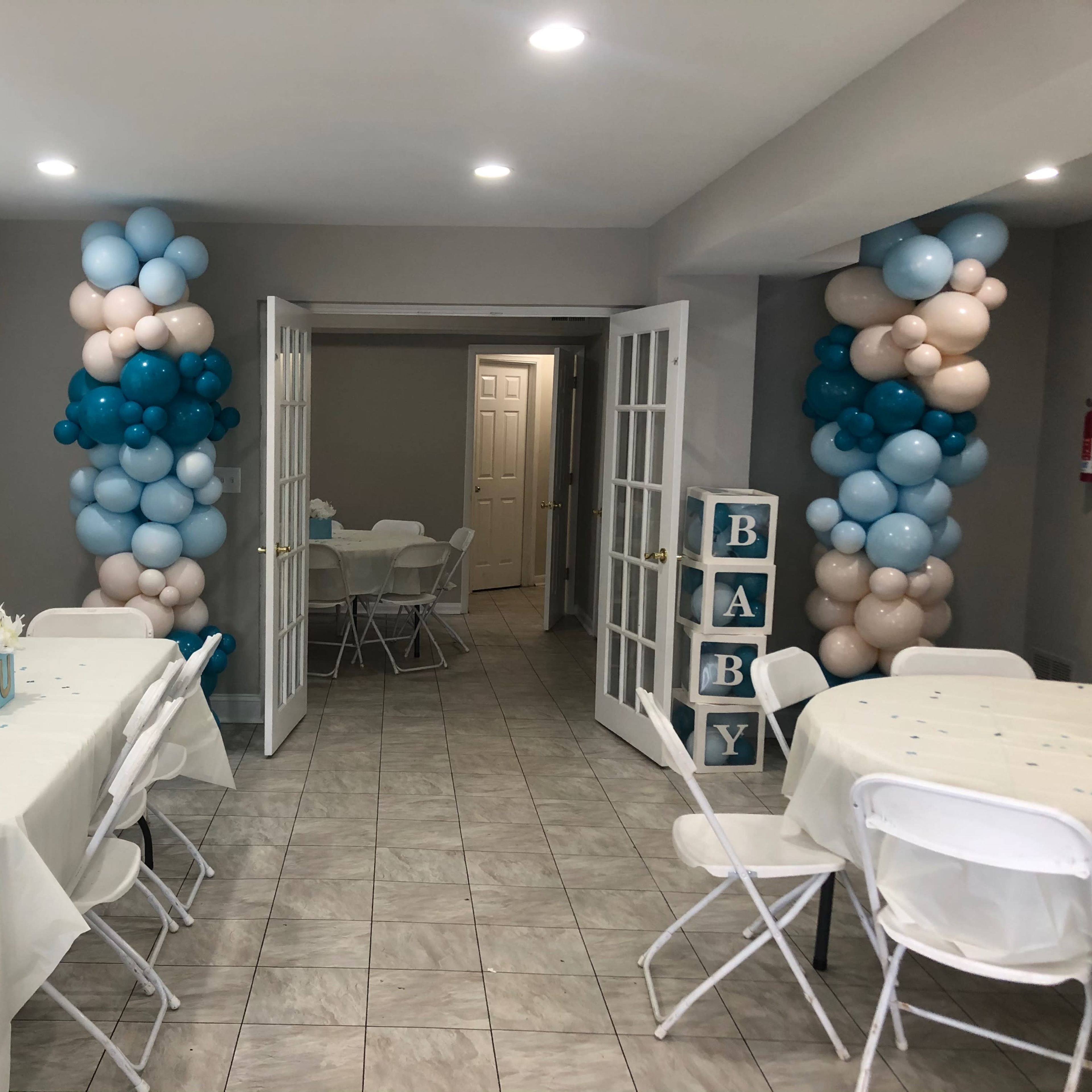 A room is decorated for a baby shower, featuring tables with light-colored tablecloths and balloon columns in blue and pink.
