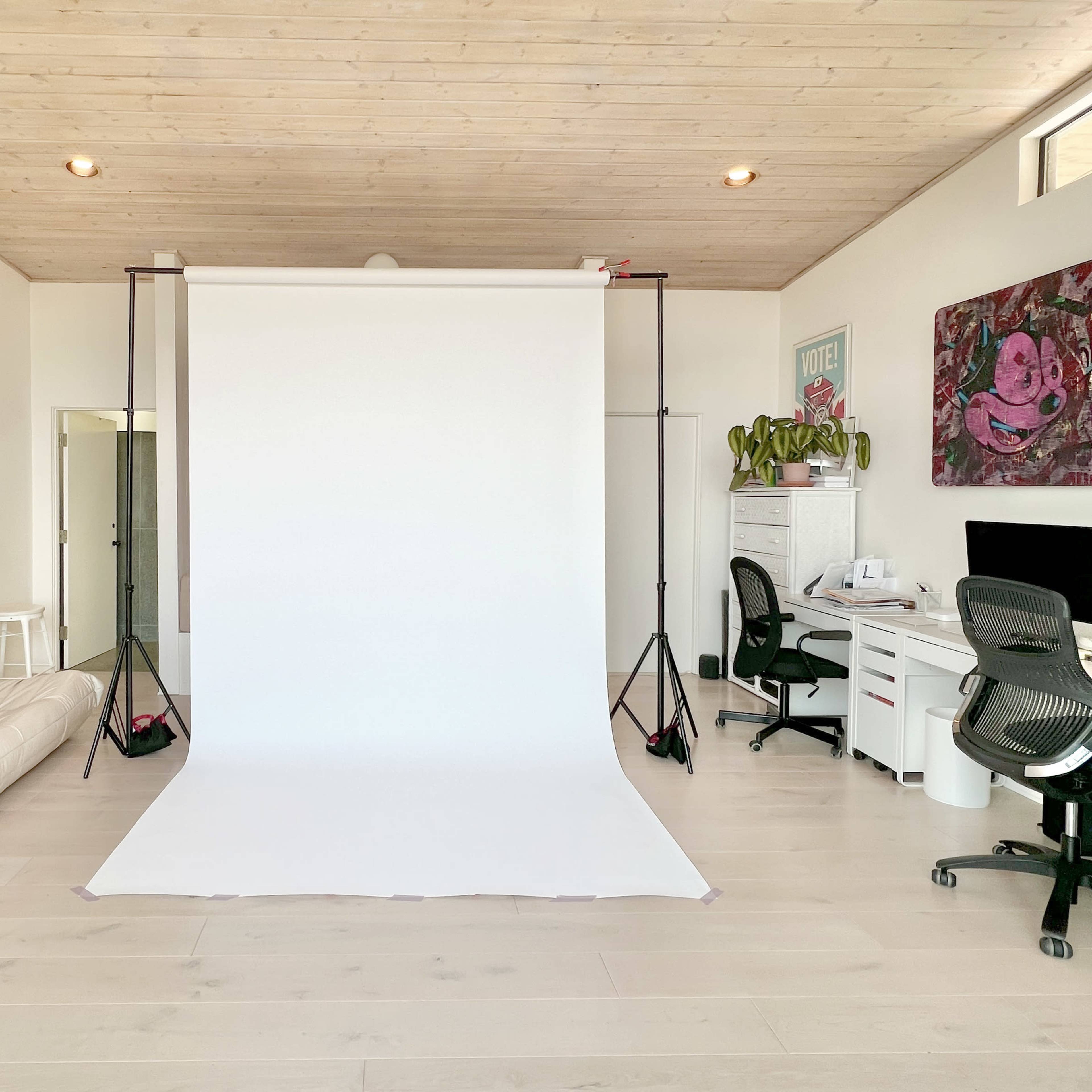 A bright, minimalist studio featuring a white backdrop on a support frame, with a sofa, desk, and decorative plants in the background.