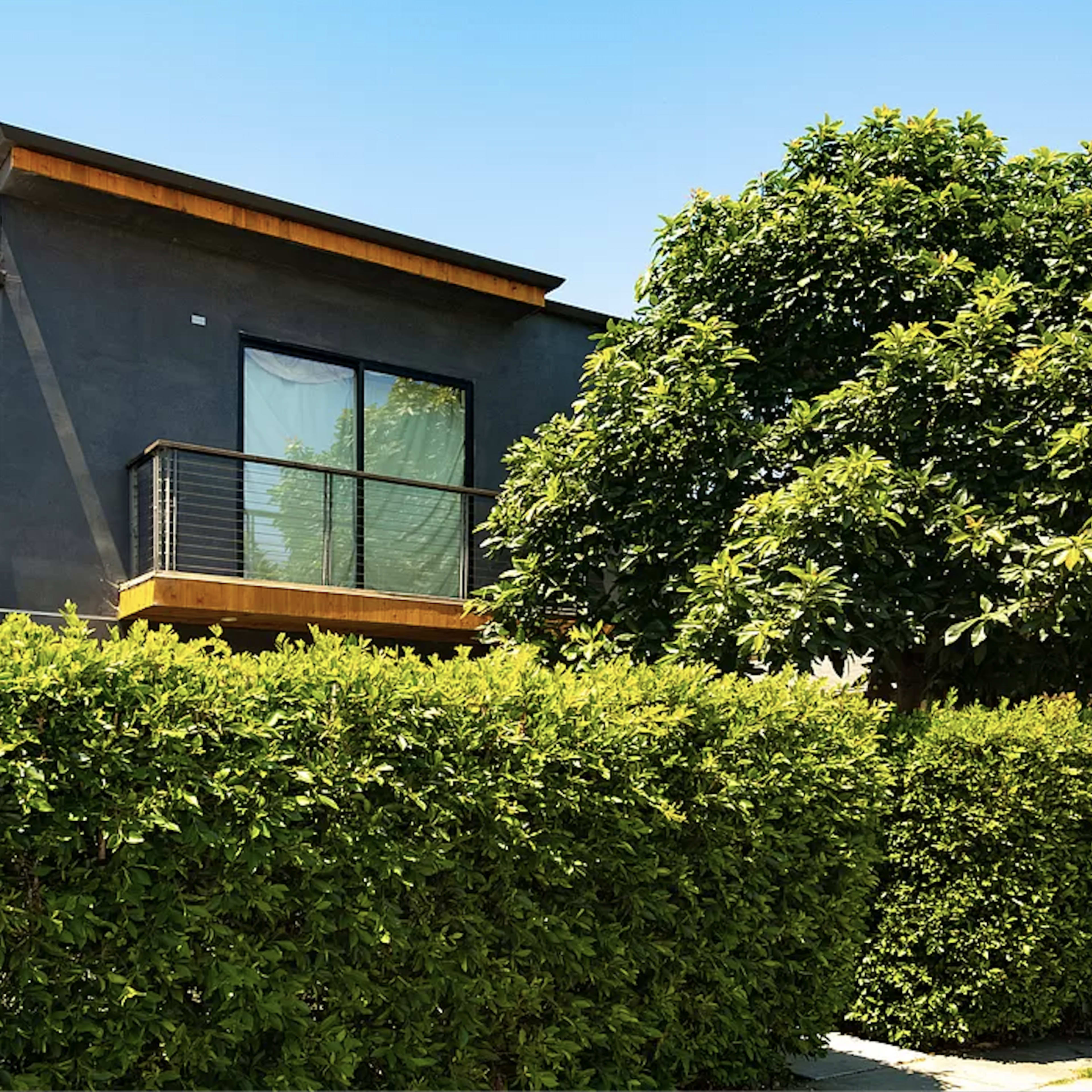 A modern two-story house with a balcony, surrounded by a neatly trimmed hedge and lush greenery.