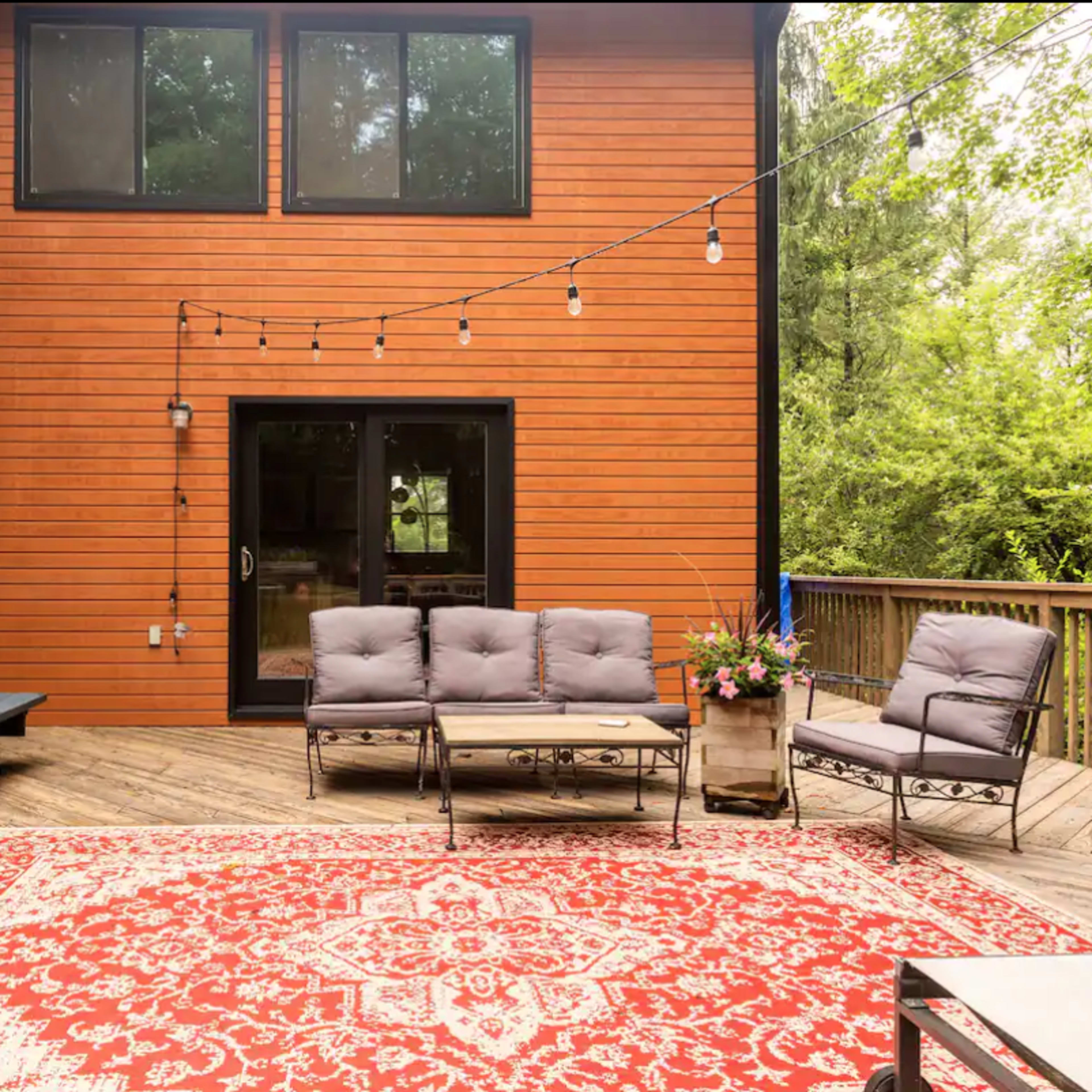A wooden deck features a seating area with a sofa and chairs, a picnic table, and a large rug, with string lights overhead and greenery in the background.