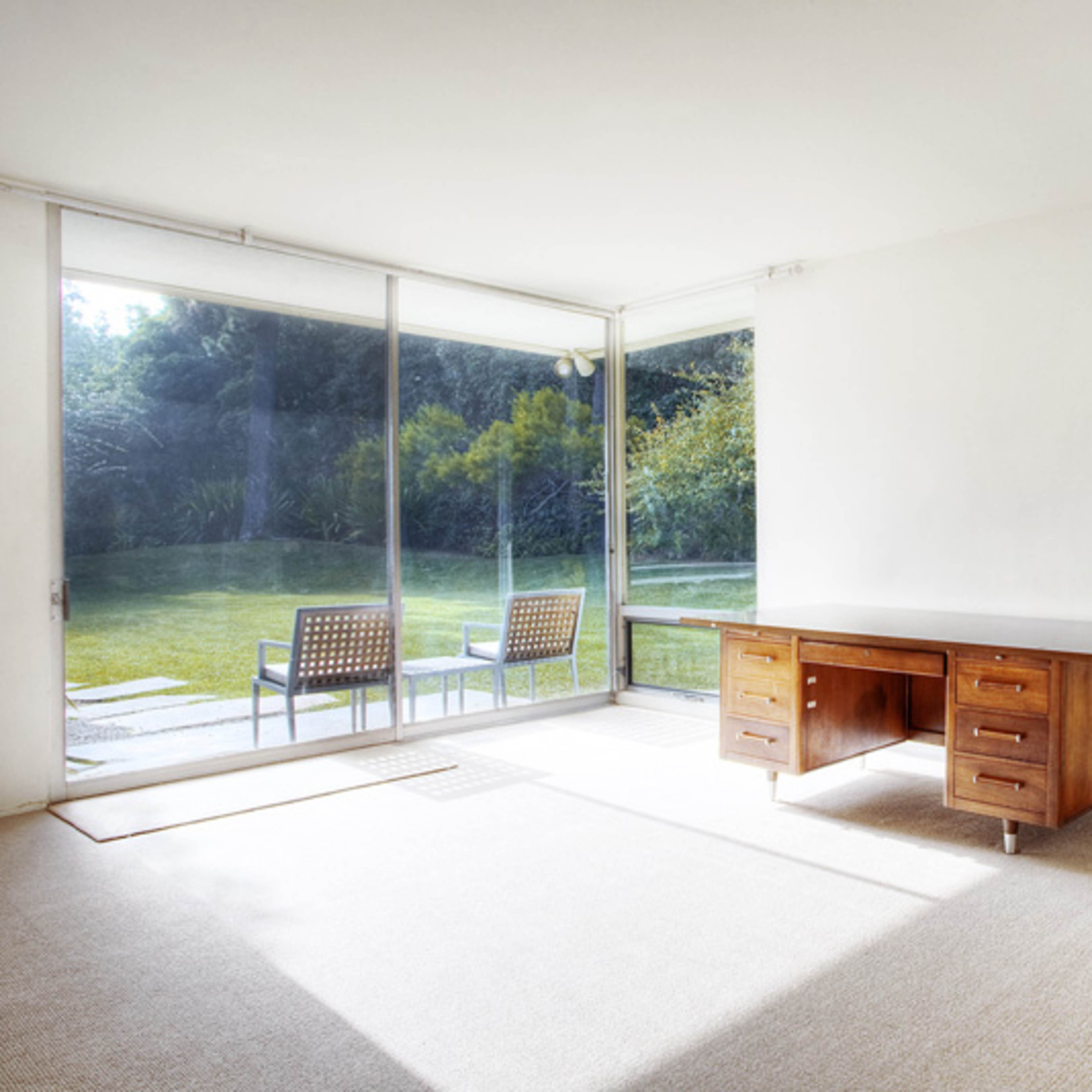 A sparsely furnished room with a wooden desk and large glass windows overlooking a manicured outdoor space.
