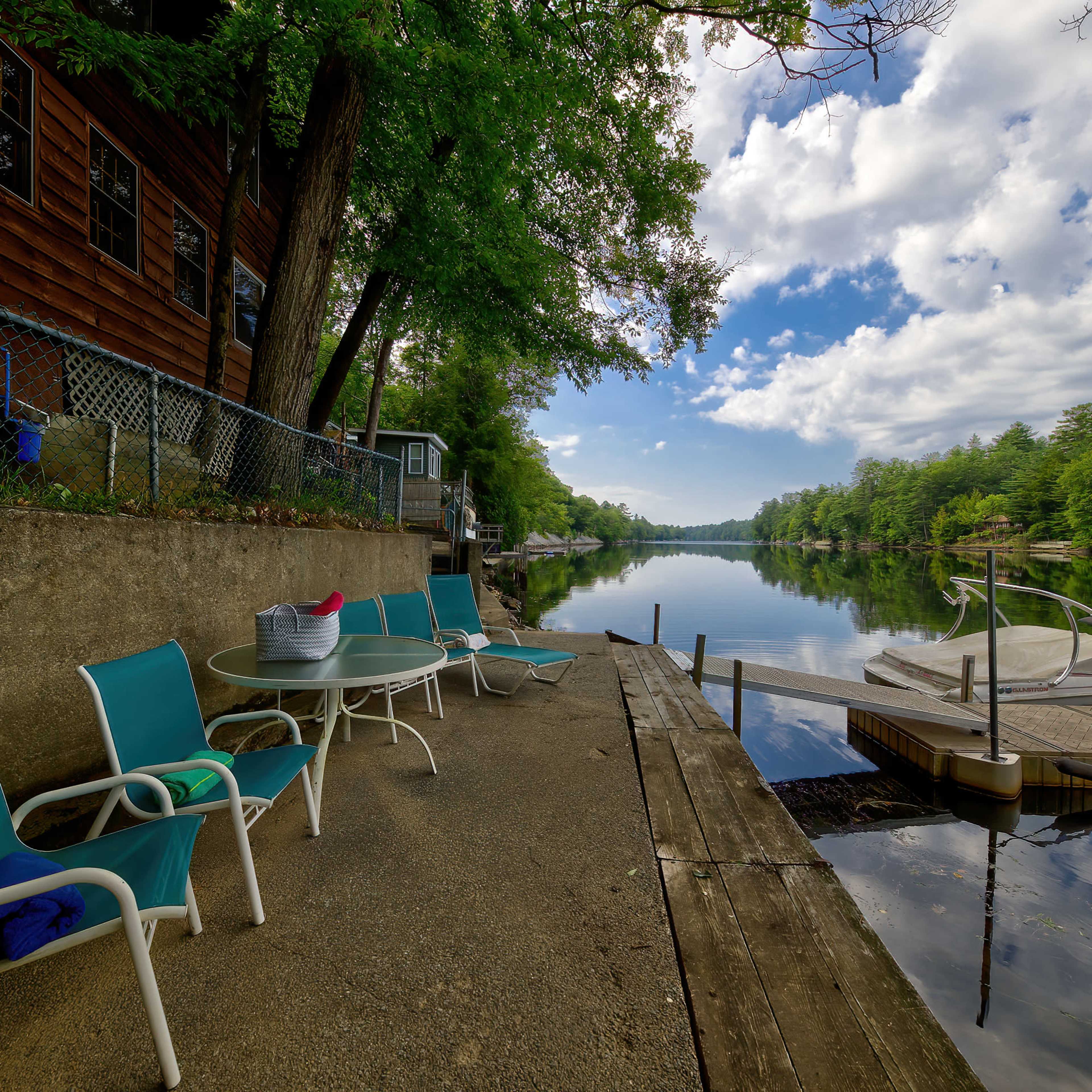 A serene lakeside scene features a wooden dock, patio chairs, and a boat tied up near a forested shoreline under a partly cloudy sky.