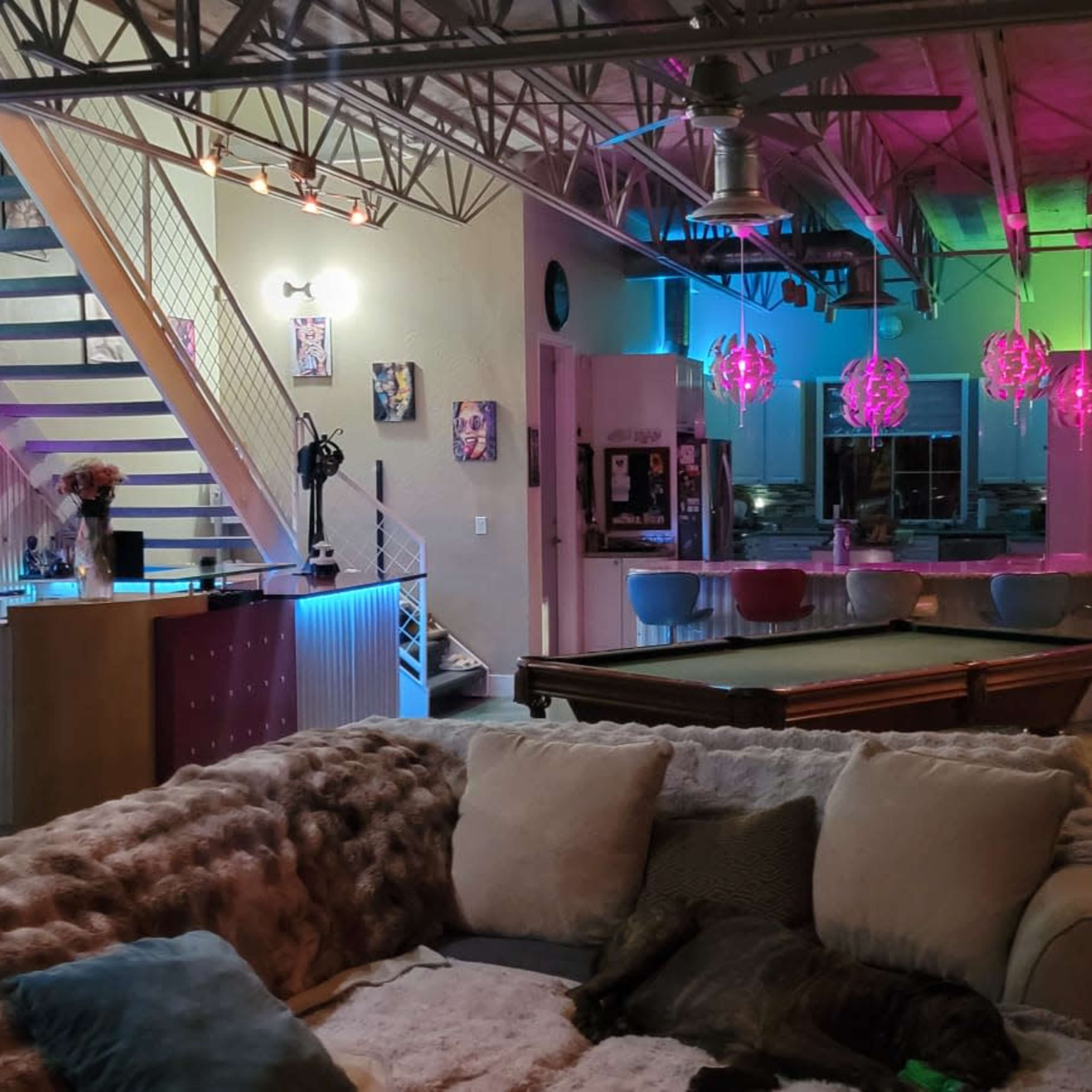 A spacious loft-style living area with a pool table, a large sectional sofa, and colorful lighting from overhead fixtures.