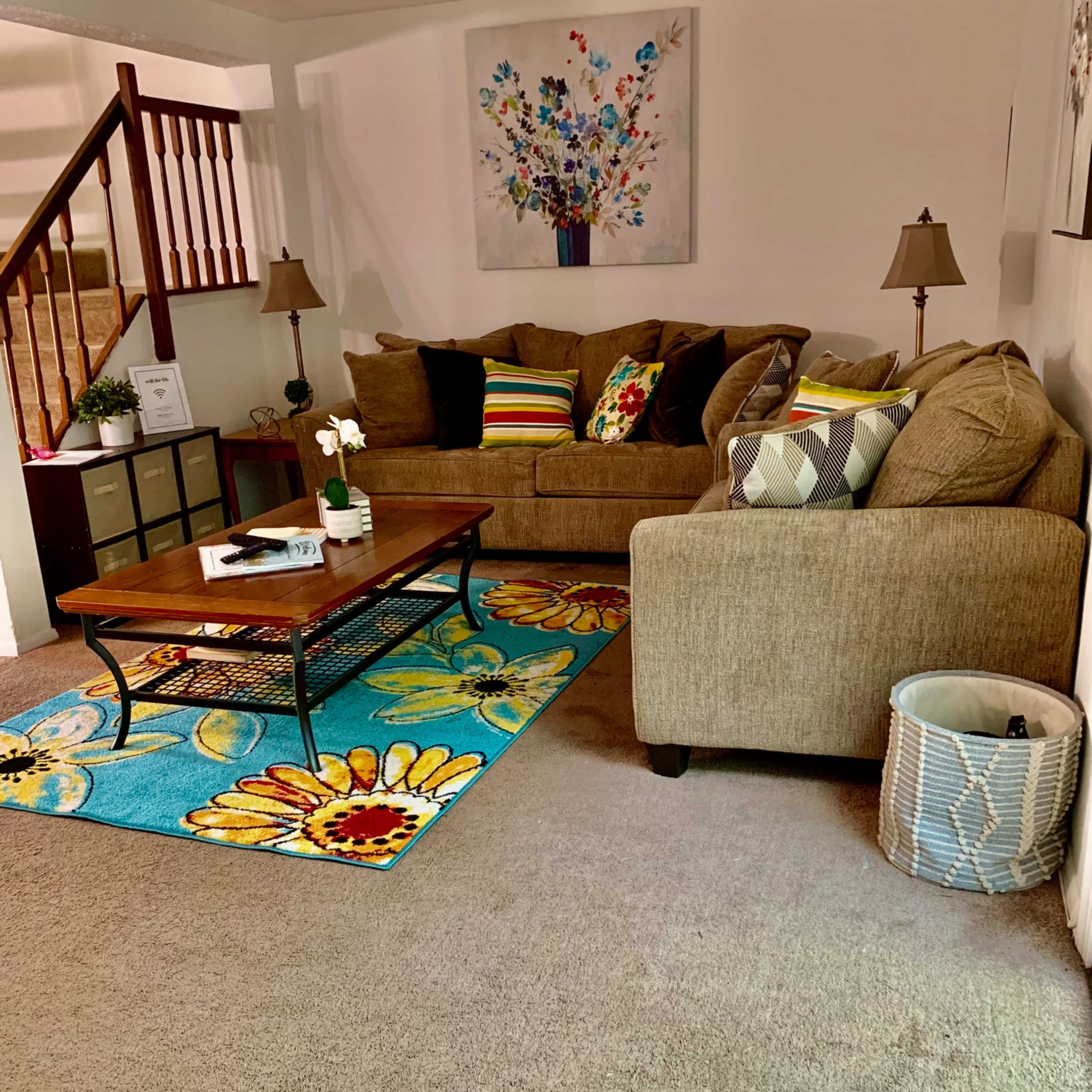 The living room features a brown couch with colorful cushions, a wooden coffee table on a bright floral rug, and a lamp beside the seating area.