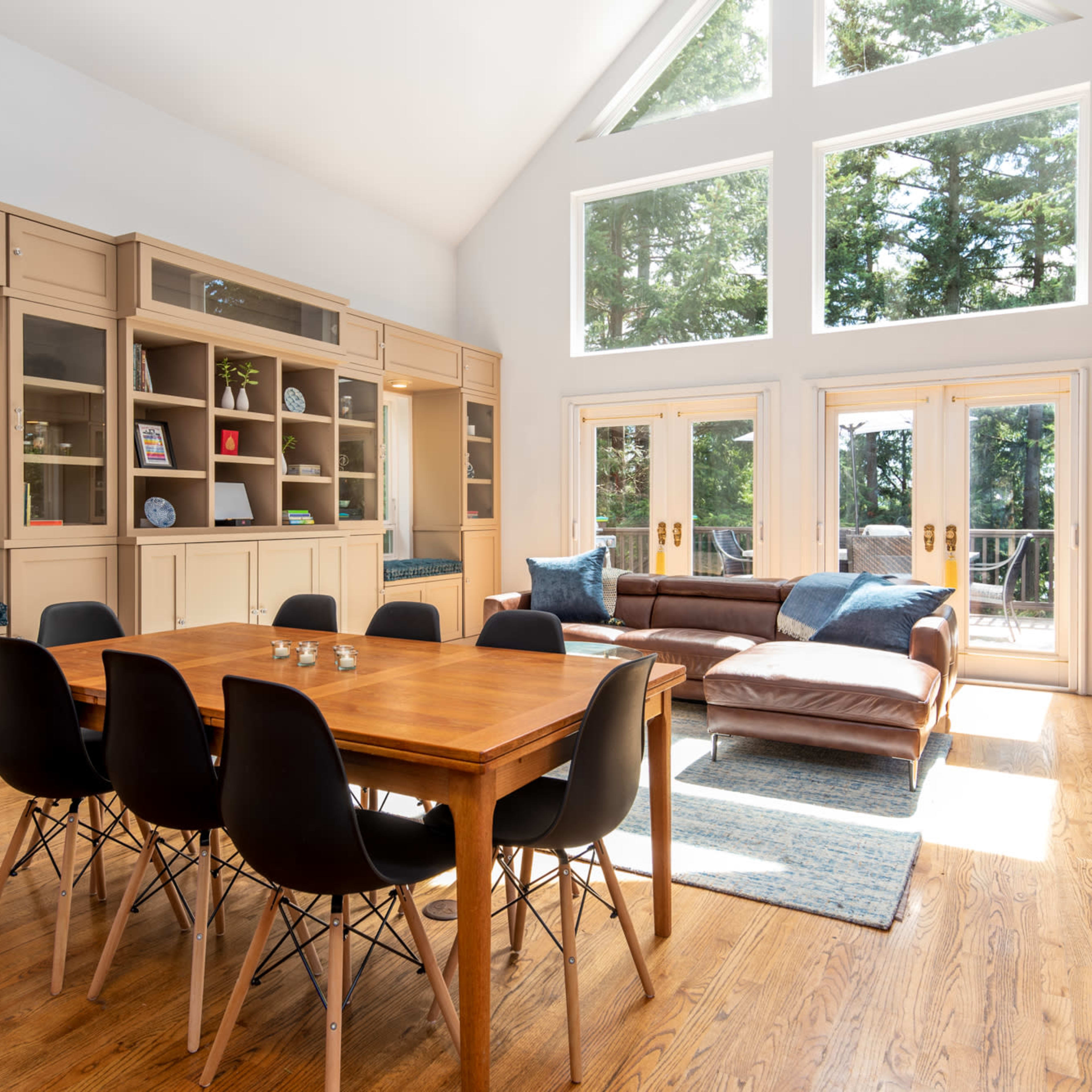A bright living room features a wooden dining table surrounded by six black chairs, with large windows allowing natural light and views of the trees outside.