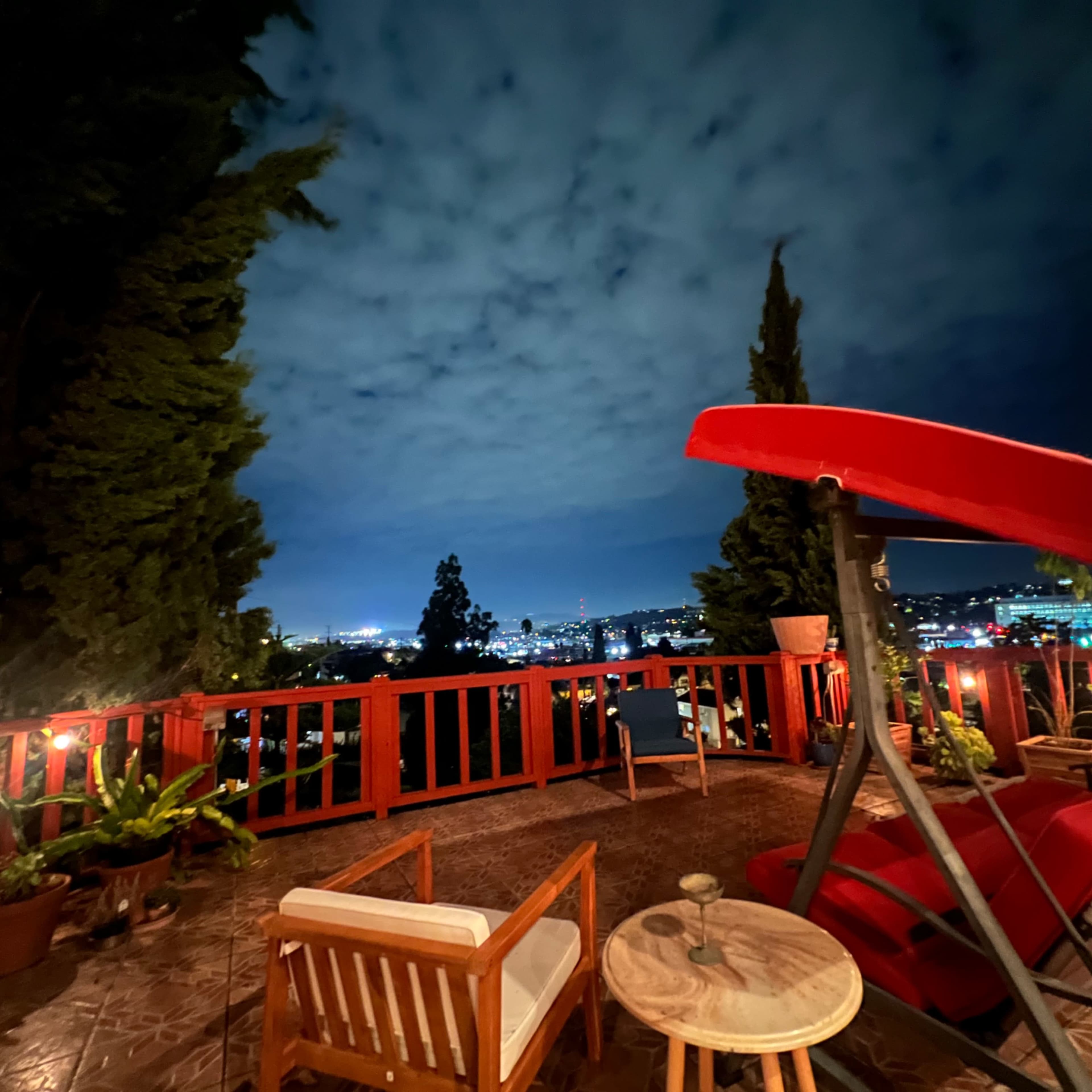 A terrace with wooden furniture and a red canopy swing overlooks a city skyline under a cloudy night sky.