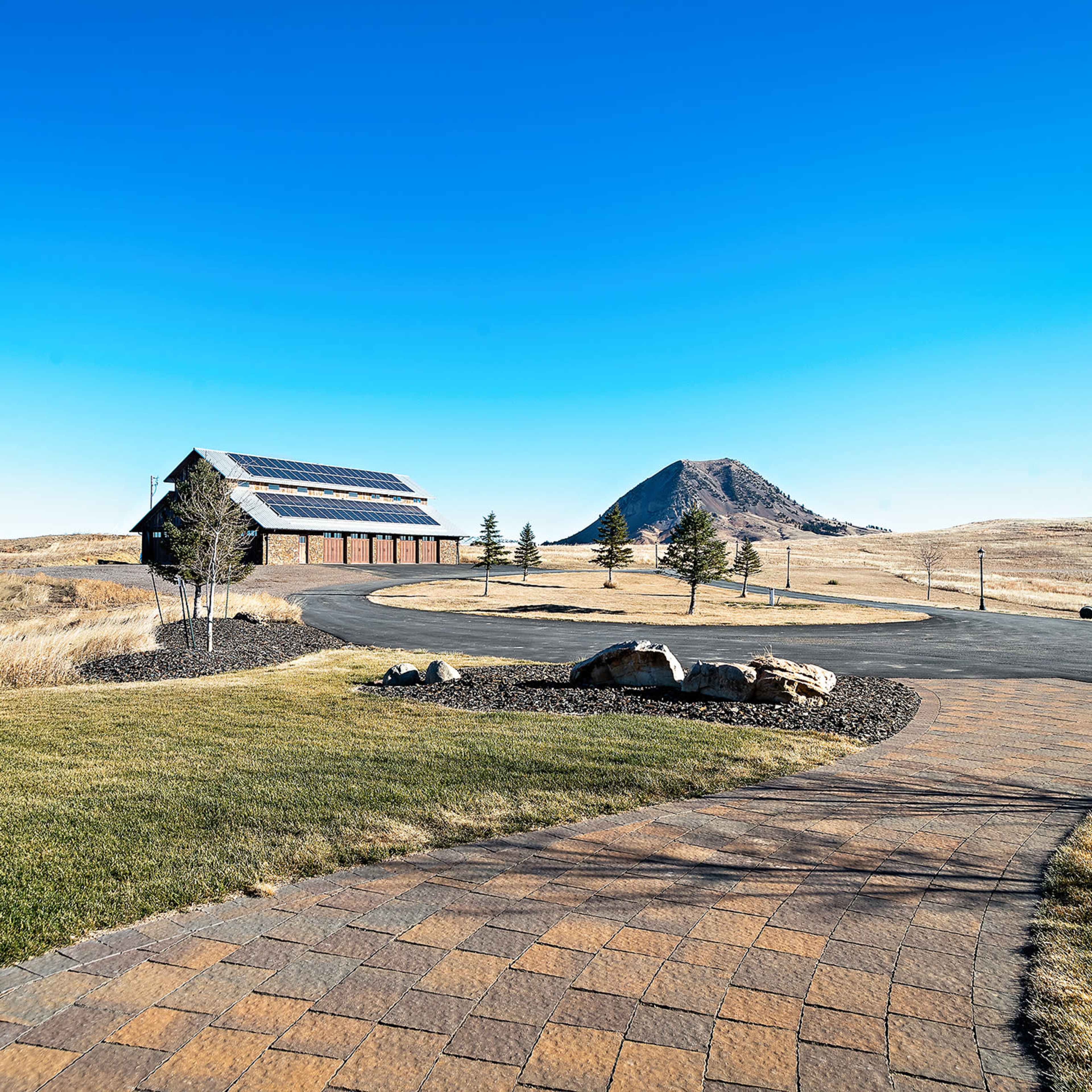 A paved pathway leads to a large barn-like structure with solar panels, set against a backdrop of rolling hills and a prominent conical hill.