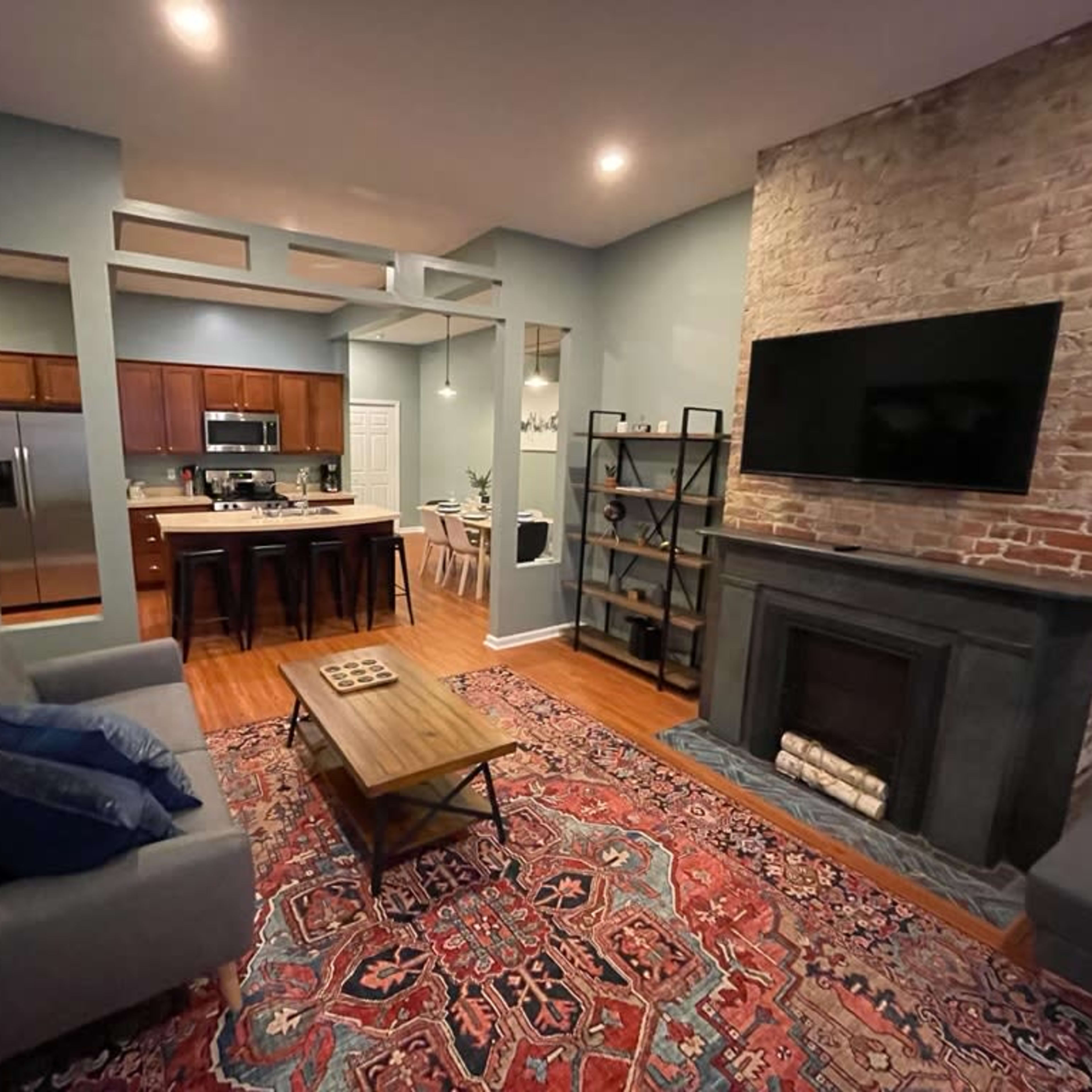A contemporary living room with a brick fireplace, a gray sofa, a wooden coffee table, and a wall-mounted TV, accompanied by a kitchen area in the background.