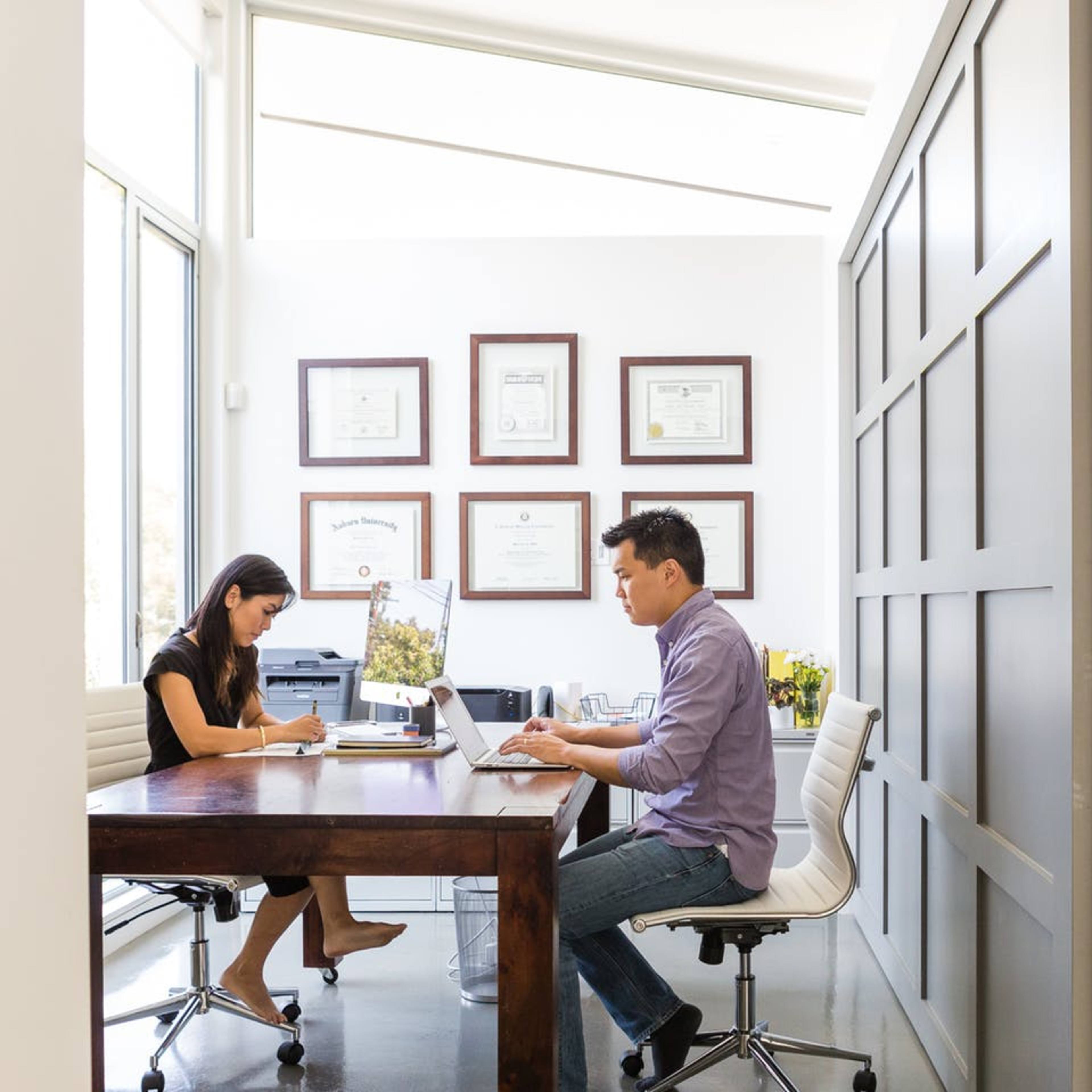 A woman and a man are seated at a wooden table in an office, working on their laptops while surrounded by framed certificates on the wall.