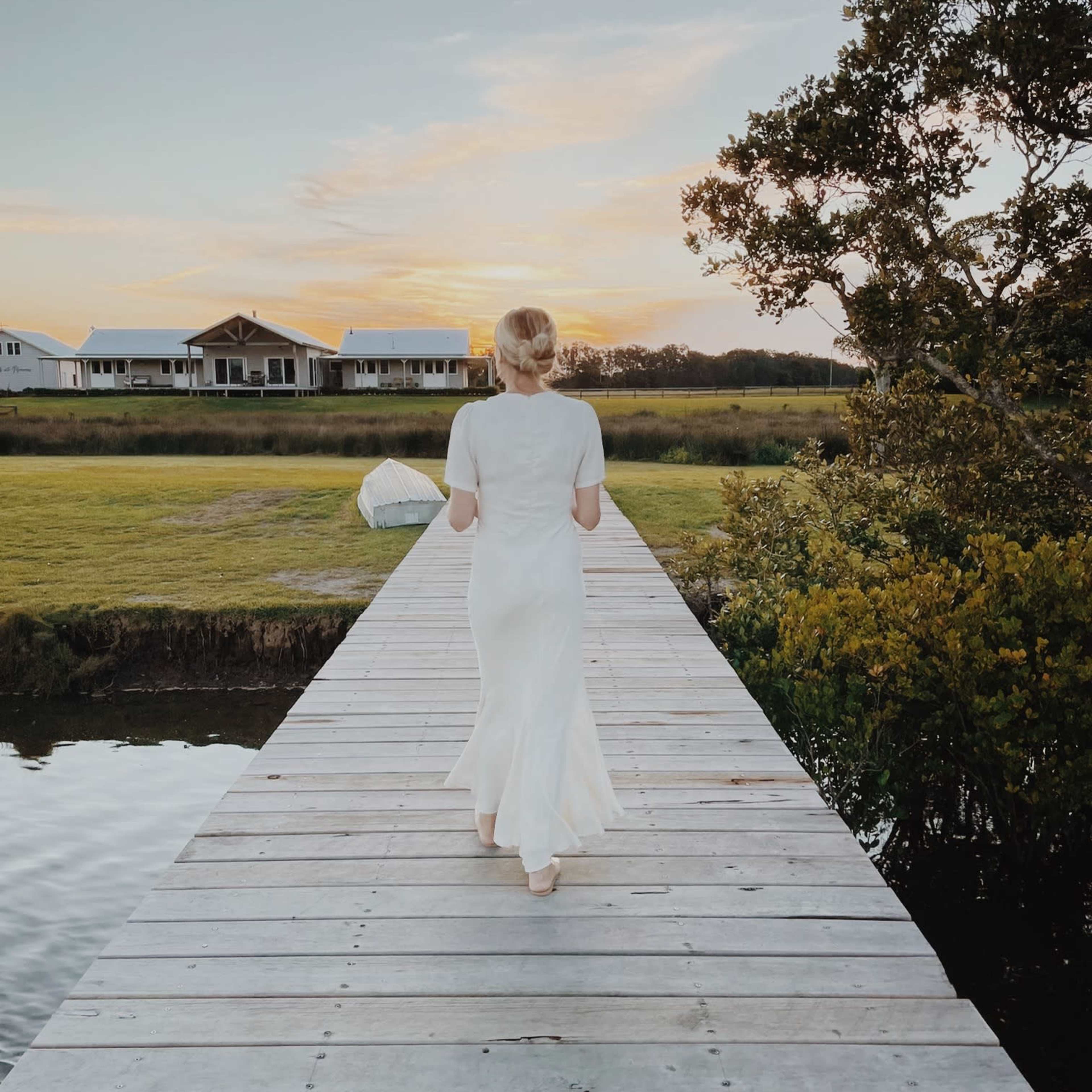 A woman in a white dress walks along a wooden dock toward a waterway at sunset.