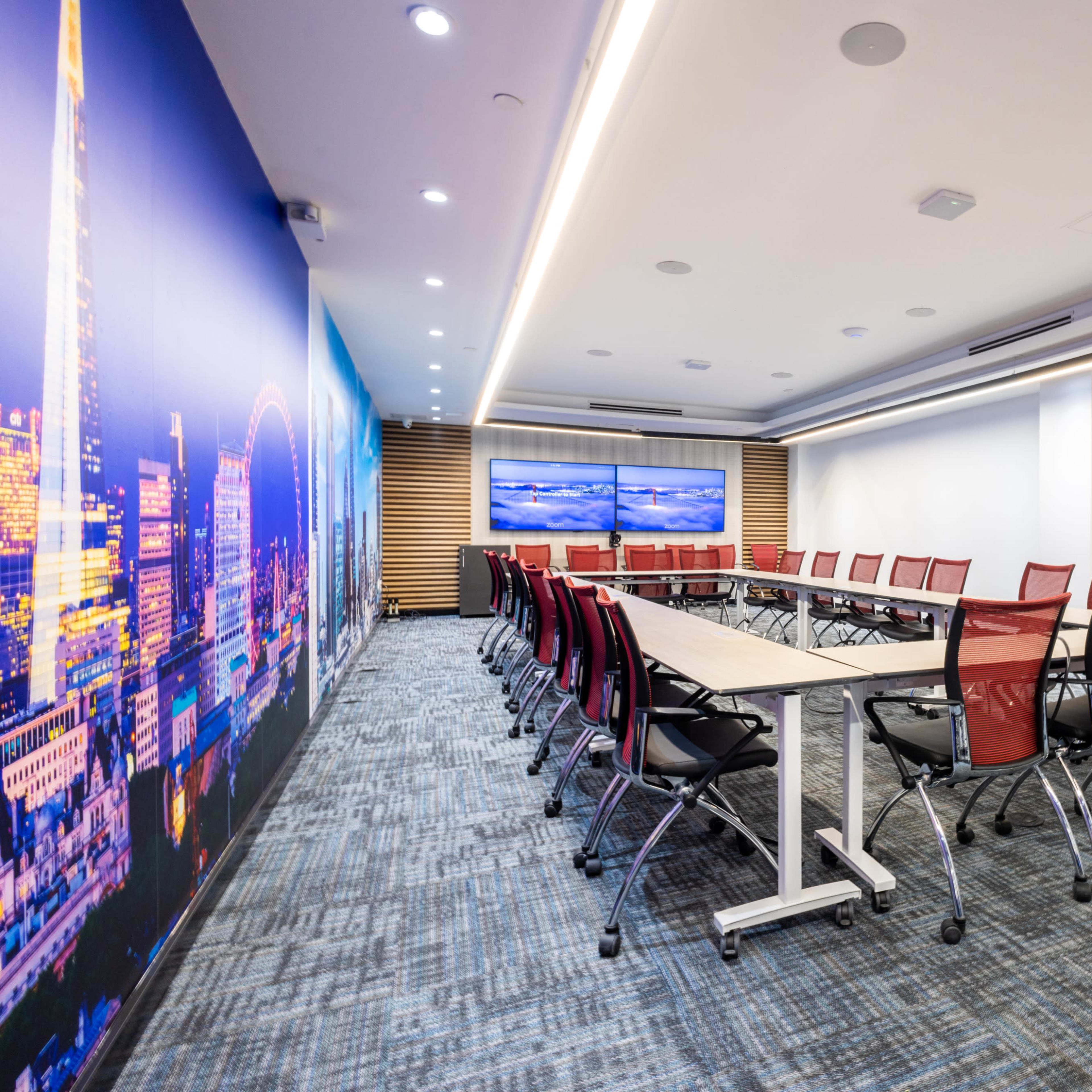 A modern conference room with a long table surrounded by red chairs, featuring large screens on the wall and a cityscape mural.