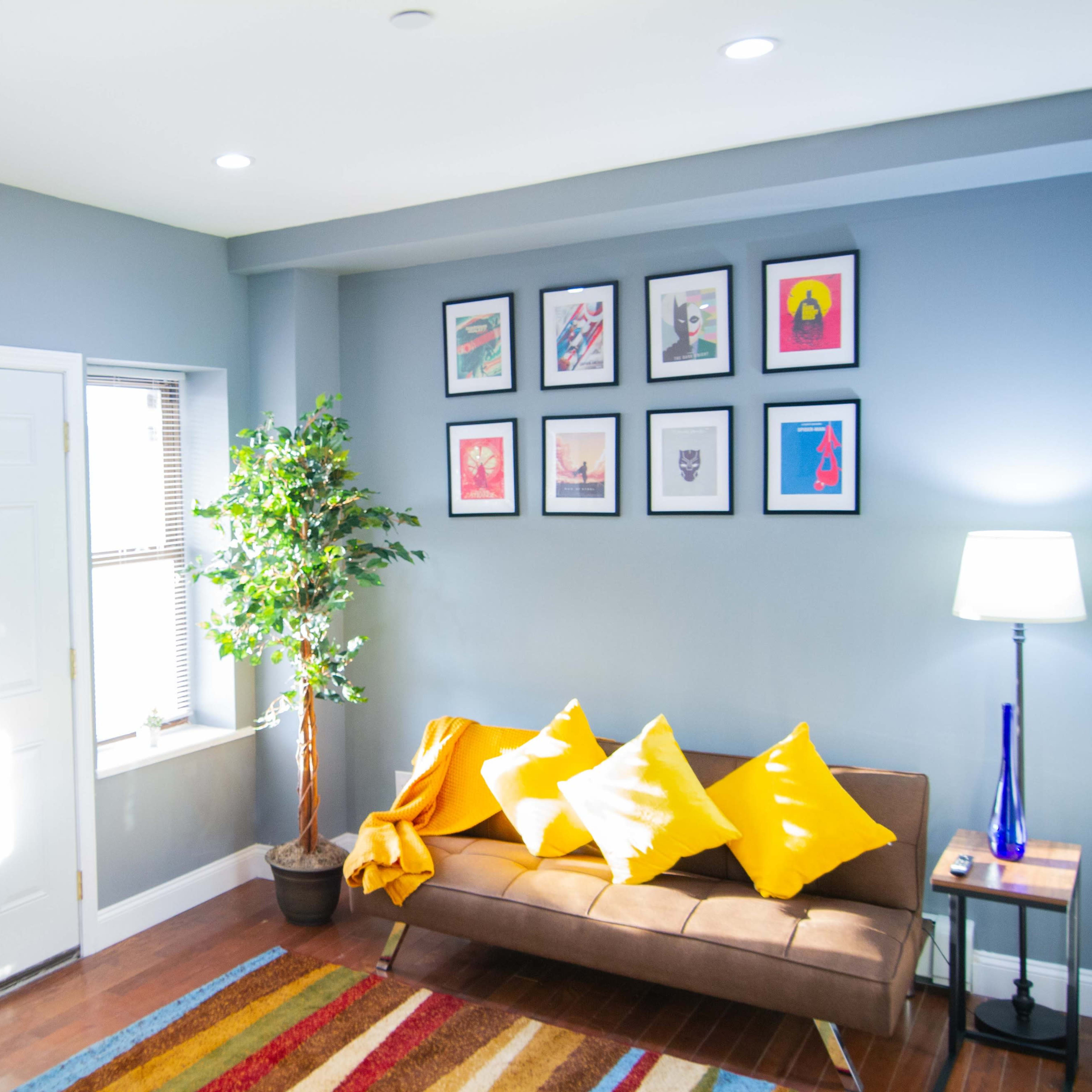 A cozy living room features a brown sofa with yellow cushions, a round dining table, colorful framed artwork on the walls, and a potted plant by the window.