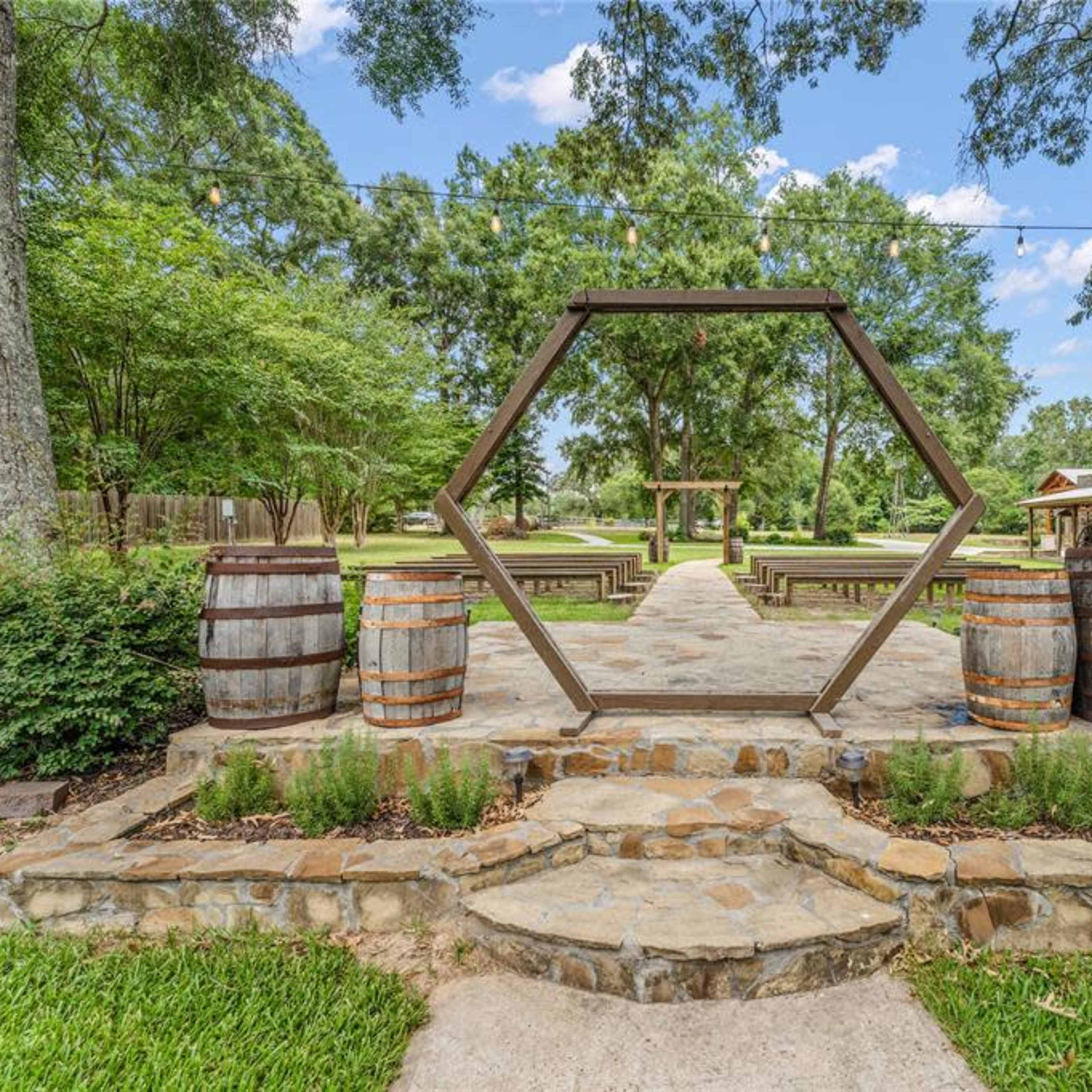 A landscaped outdoor setting with a hexagonal wooden structure at the center, surrounded by stone paths, wooden barrels, and rows of seating.