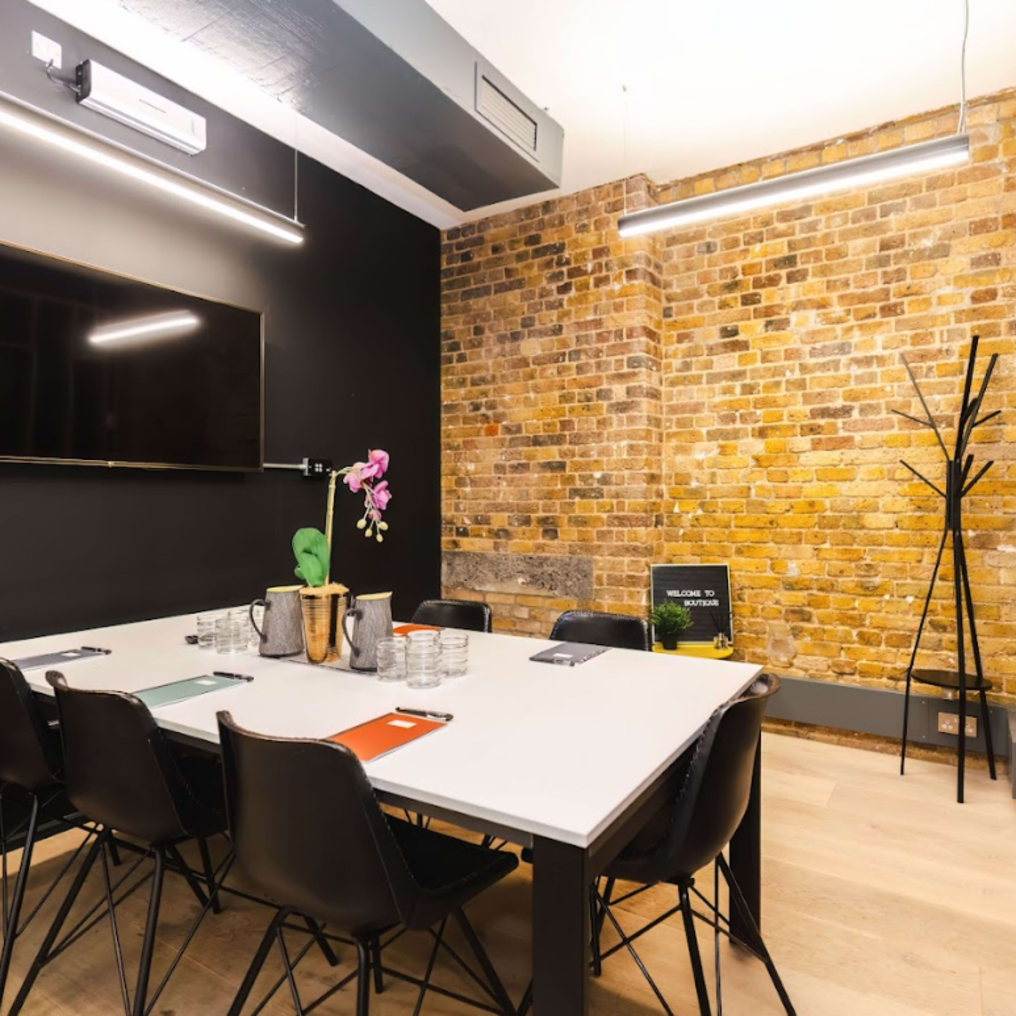 A conference room features a large table, black chairs, a wall-mounted TV, and exposed brick walls.
