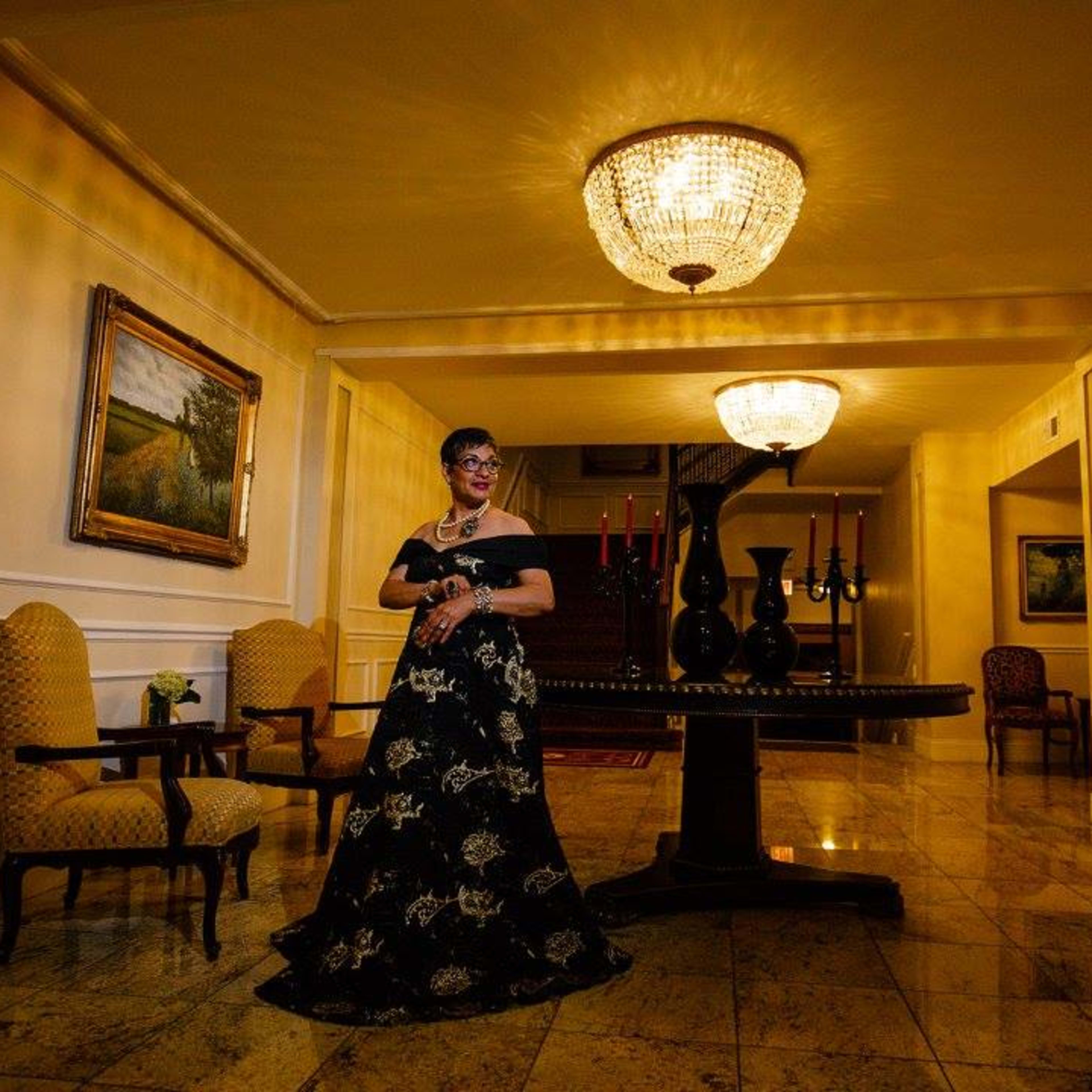 A woman in an elegant black dress stands beside a large table under a chandelier in a well-lit hallway adorned with paintings and chairs.