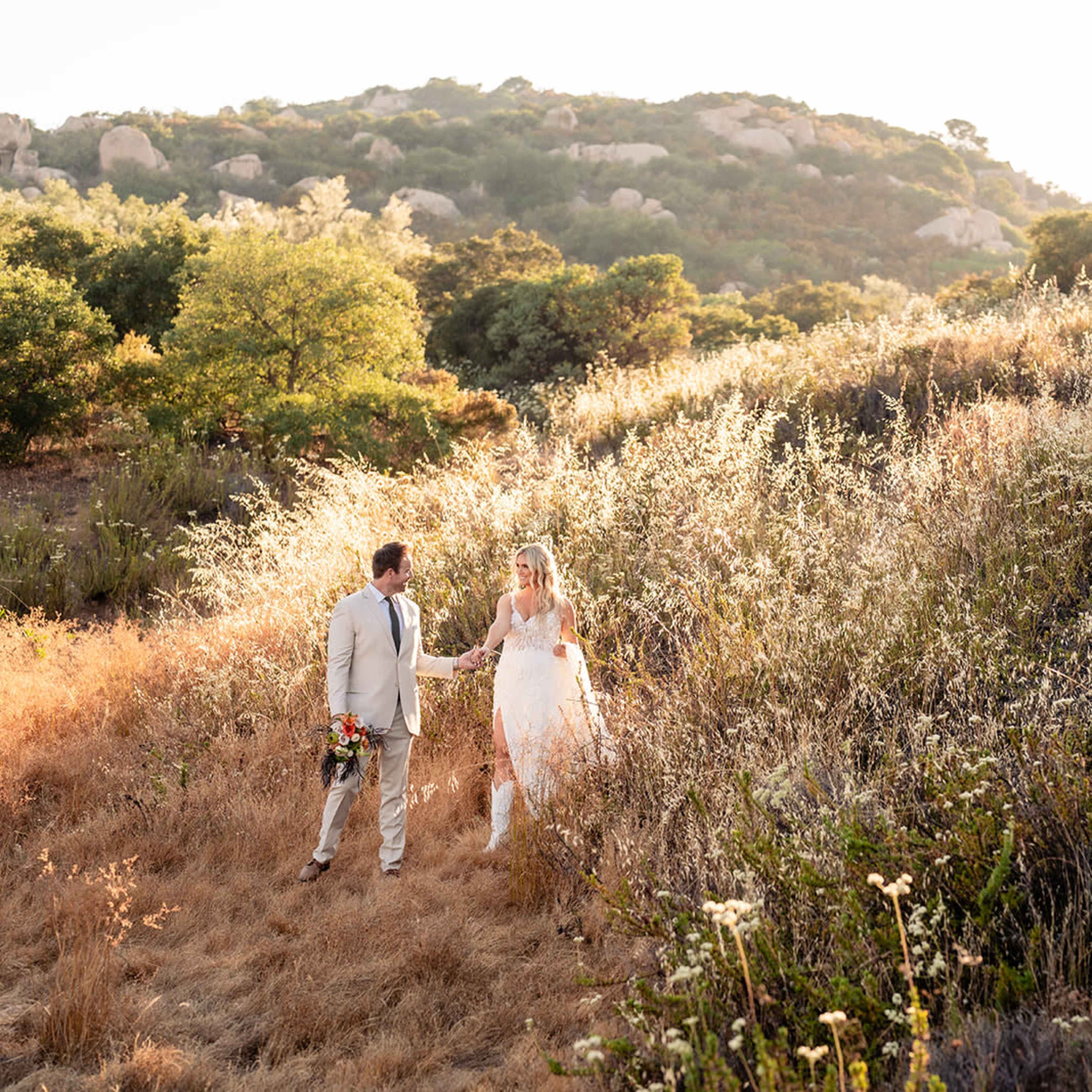 A couple dressed in wedding attire walks hand-in-hand through a sunlit, grassy landscape with scattered shrubs and trees.