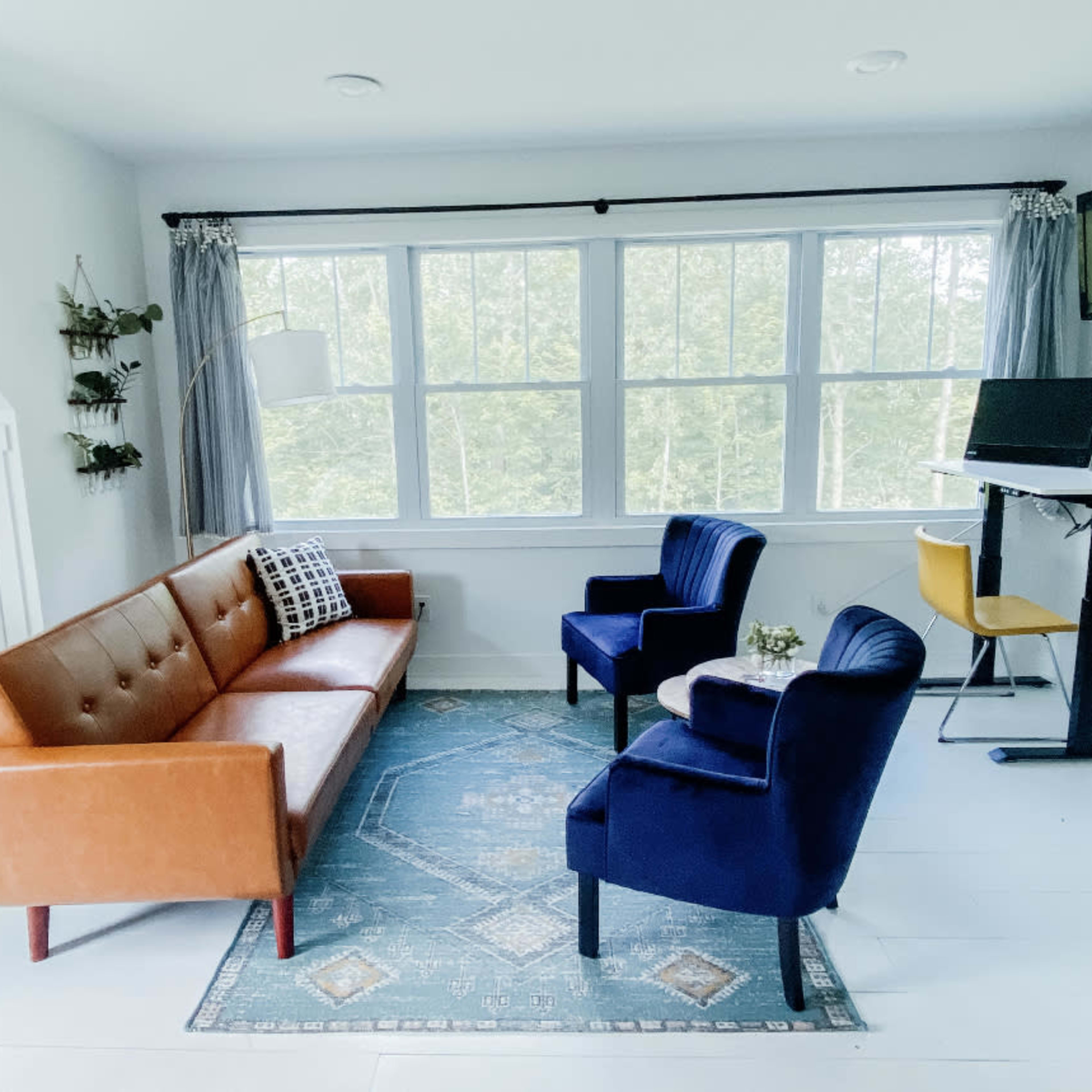 A bright room with a brown leather sofa, two blue chairs, a small table, and large windows that overlook greenery outside.