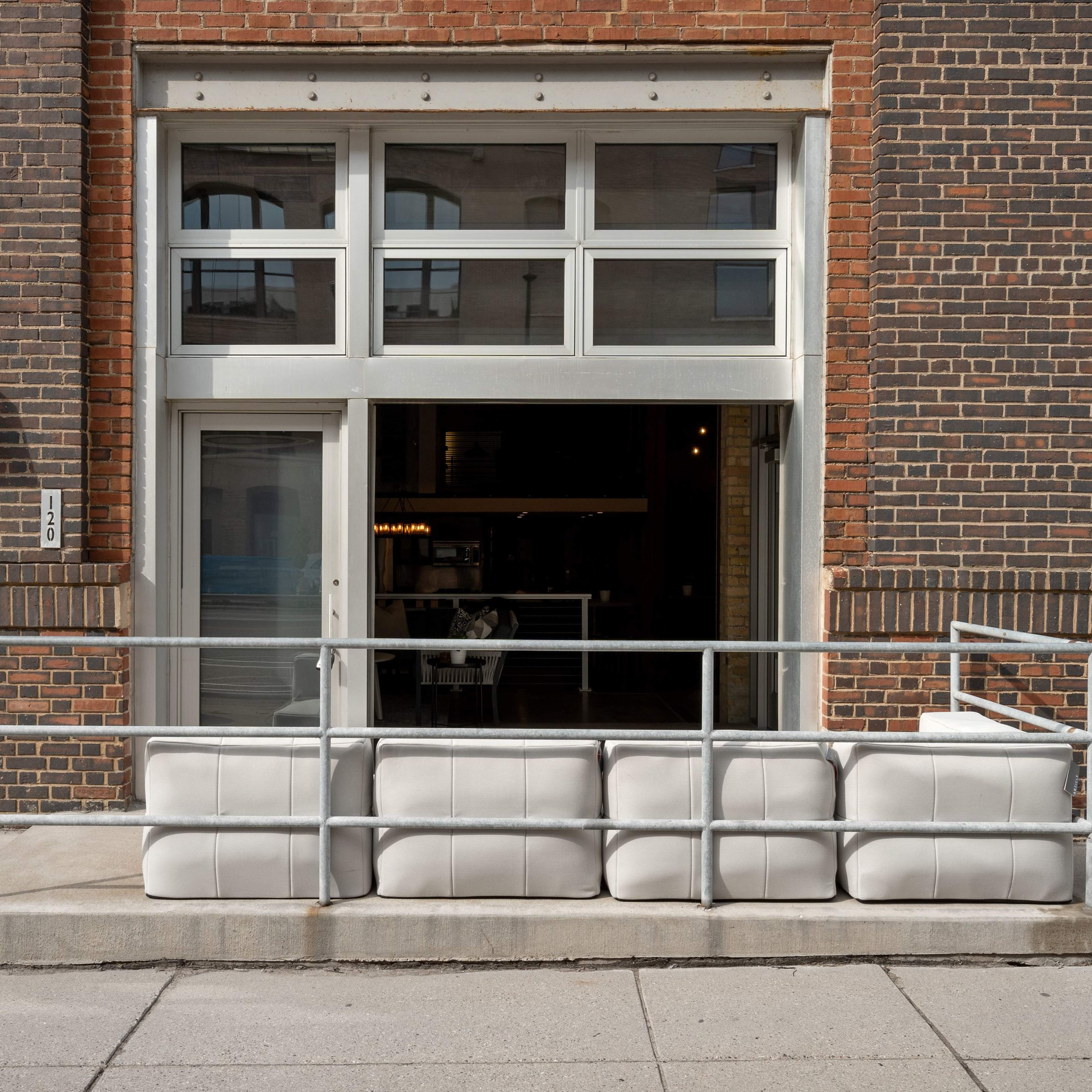 A brick building with large glass doors, featuring a railing and a row of light-colored seating elements along the entrance.