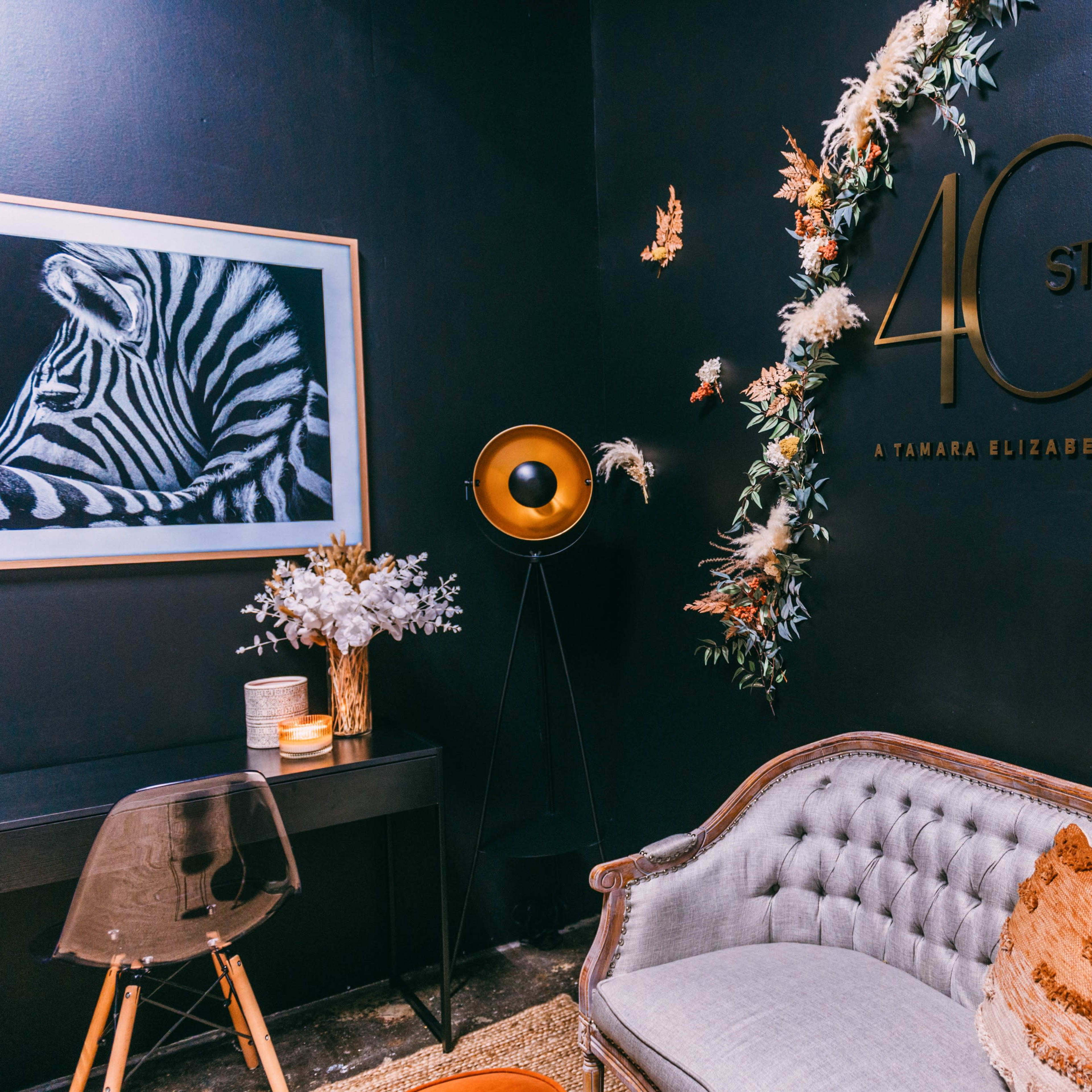 A stylish interior featuring a decorative wall with a sign that reads "40E STUDIO," a plush couch, a modern lamp, and a floral arrangement.