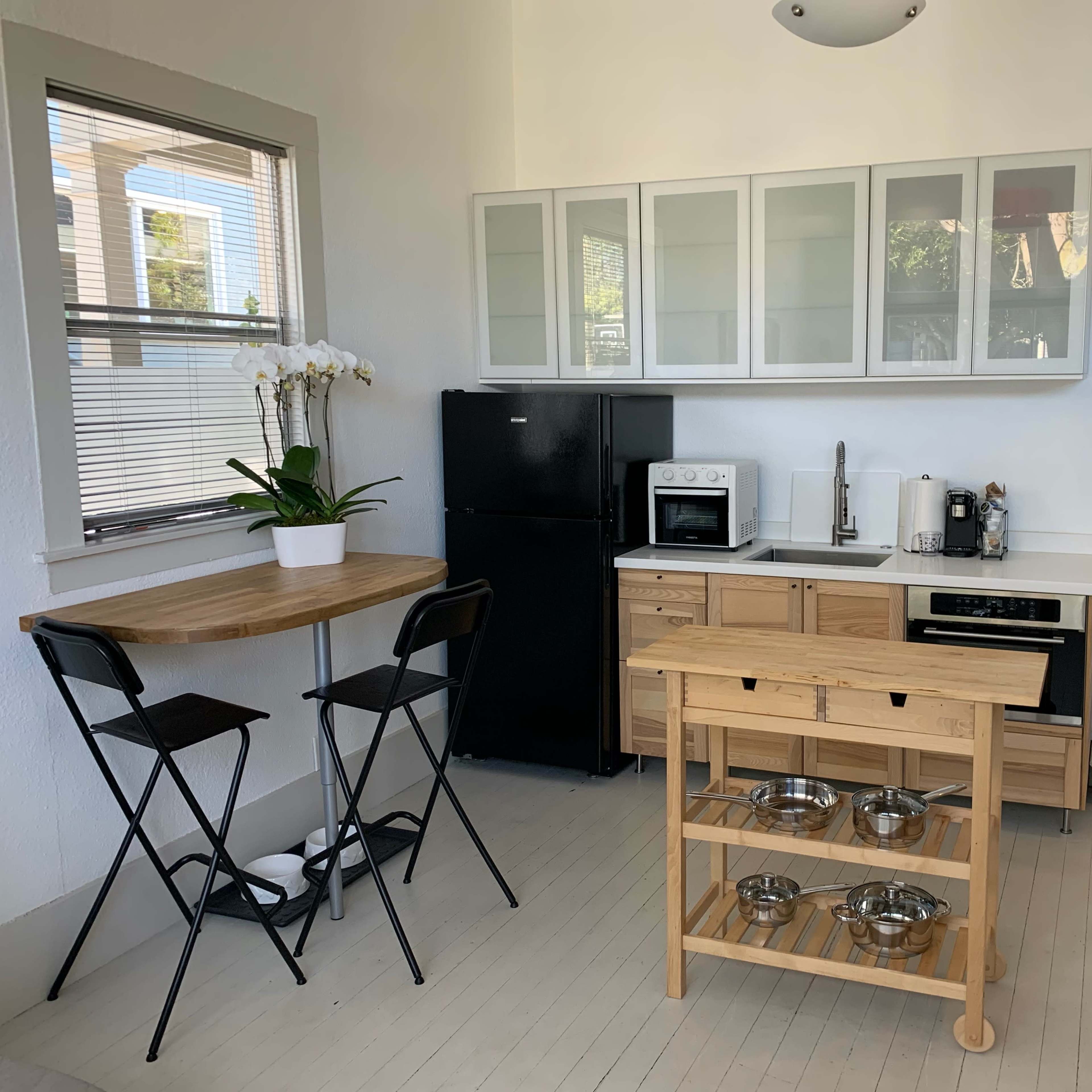 A small kitchen area featuring a black refrigerator, white cabinetry, a microwave, and a wooden table with two folding chairs.