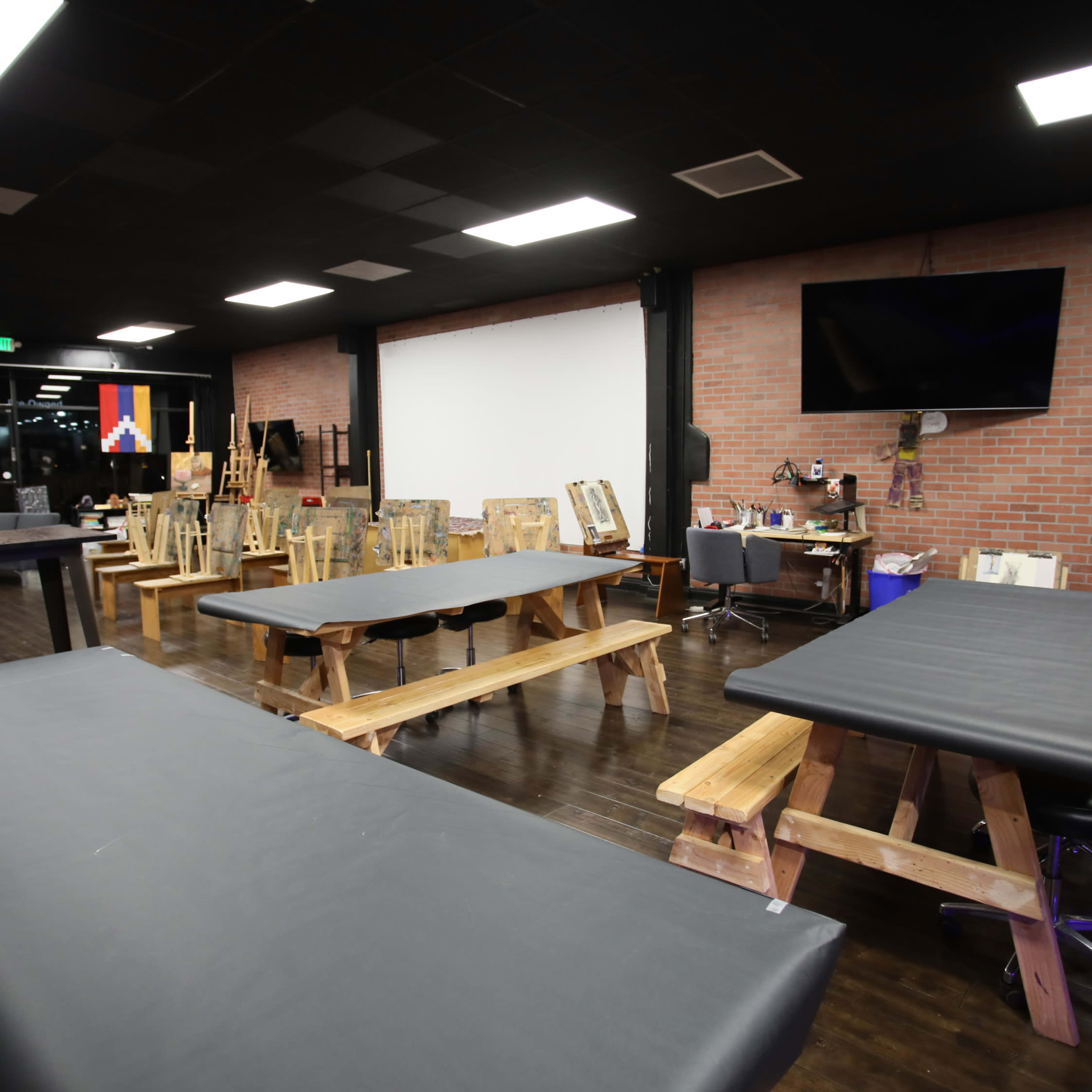 A spacious interior with several wooden tables and chairs in a studio, featuring a large blank wall, a television, and various art supplies in the background.
