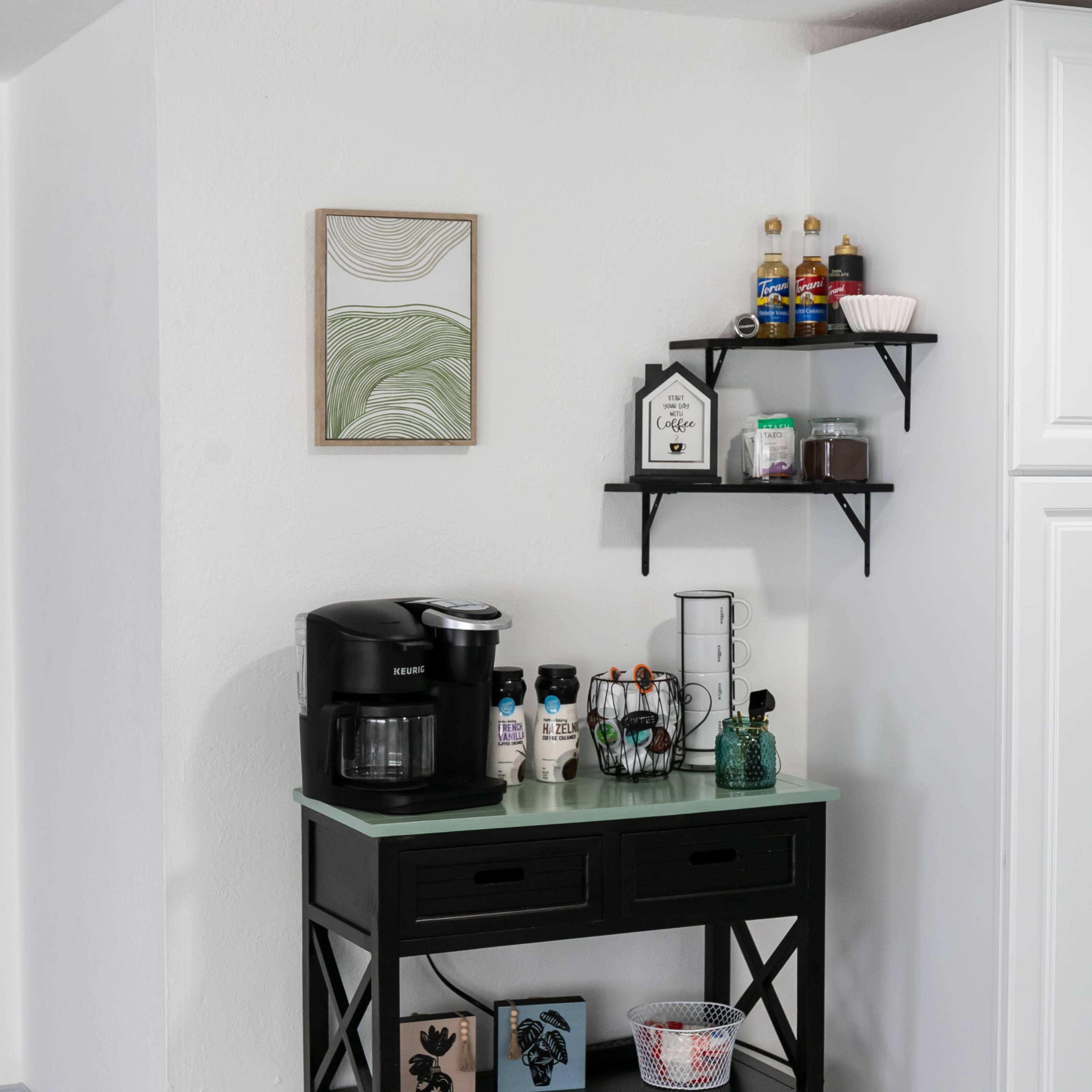 A black coffee maker is positioned on a dark console table beneath a wall-mounted shelf displaying various condiments and a small framed piece of artwork.