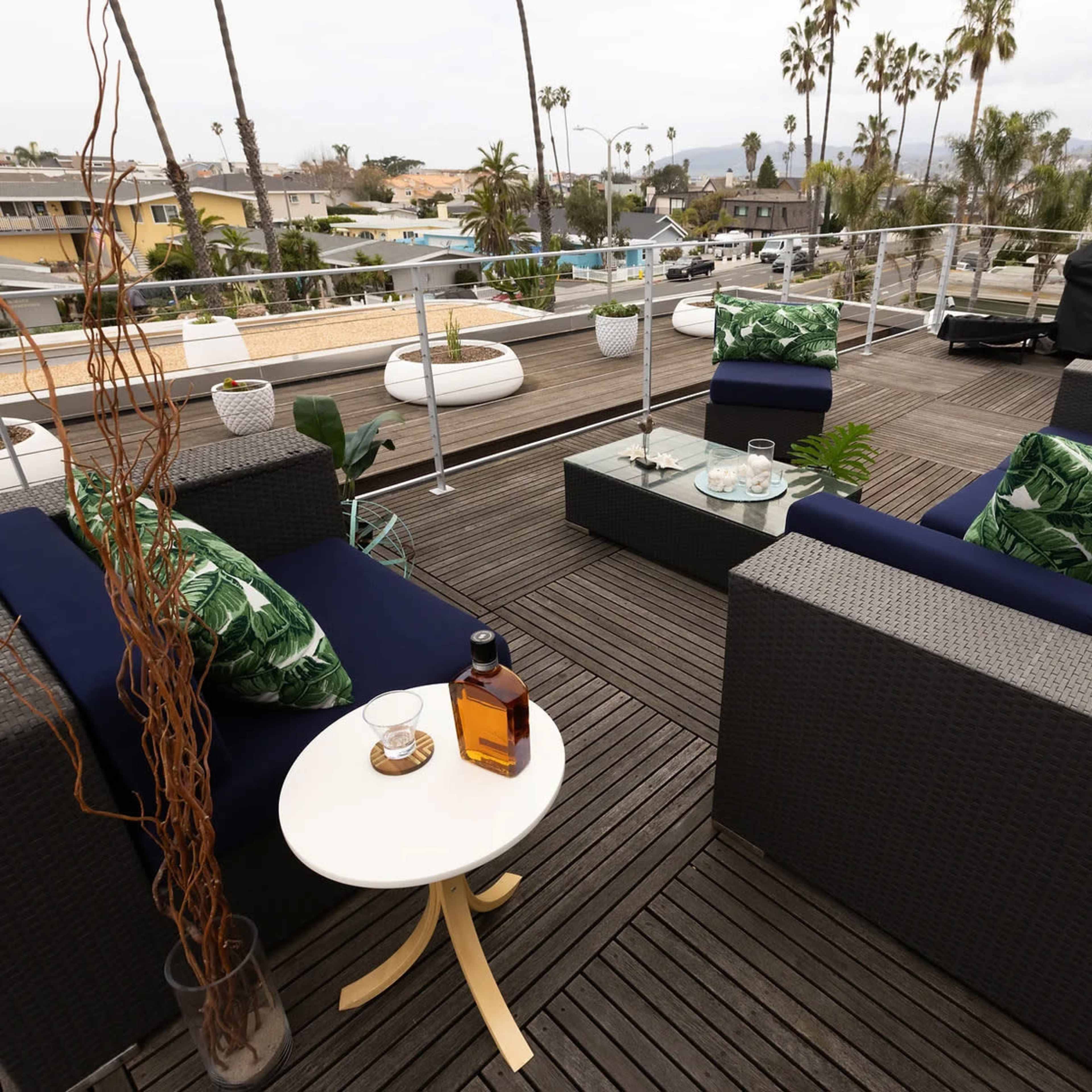 A rooftop patio features wicker seating with green leaf-patterned cushions, a glass coffee table, and a view of palm trees and buildings in the background.