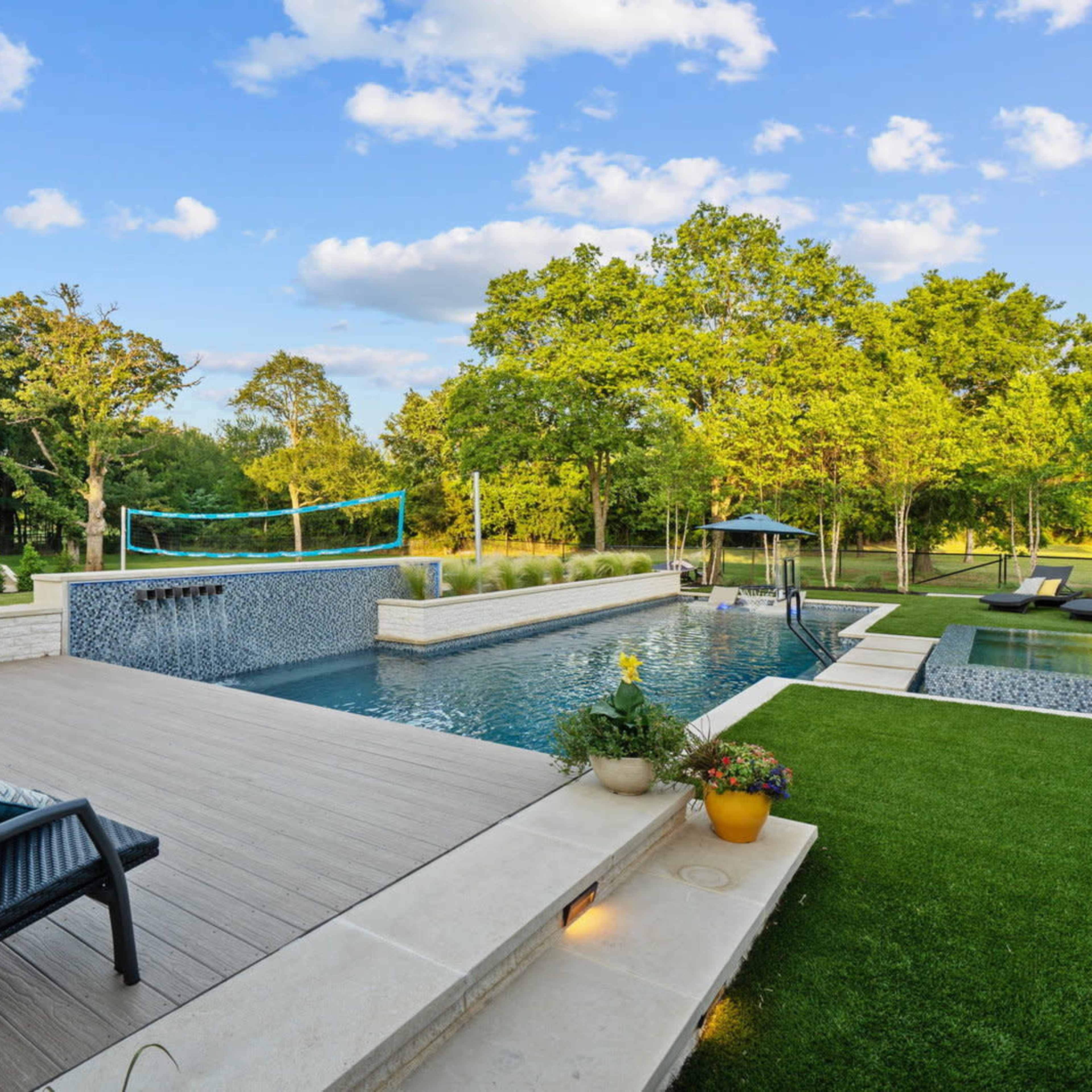 A modern outdoor space features a swimming pool surrounded by lounge chairs and a landscaped garden with trees in the background.
