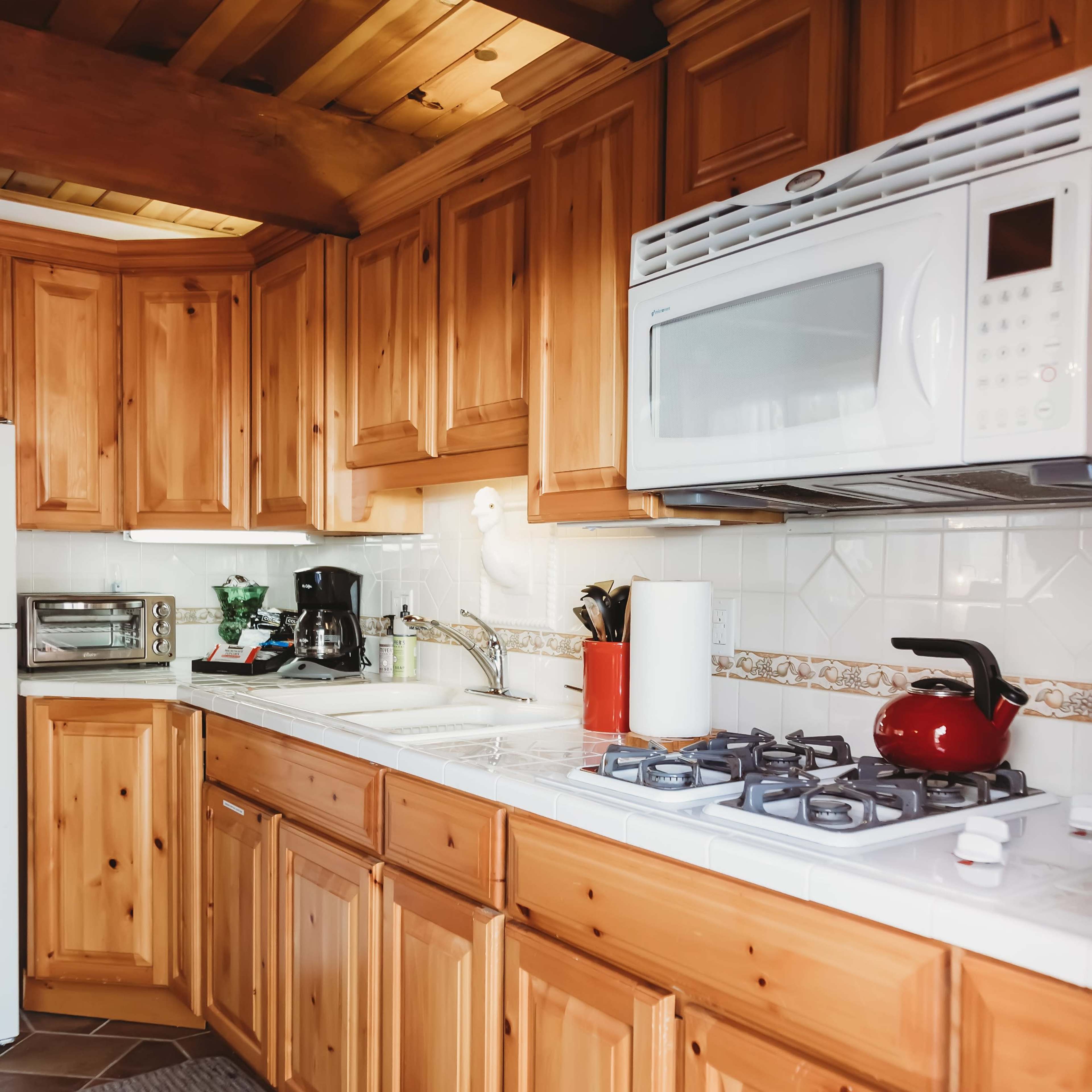 A wooden kitchen with light-colored cabinetry, a white microwave, a red kettle, and various kitchen appliances arranged along a countertop.
