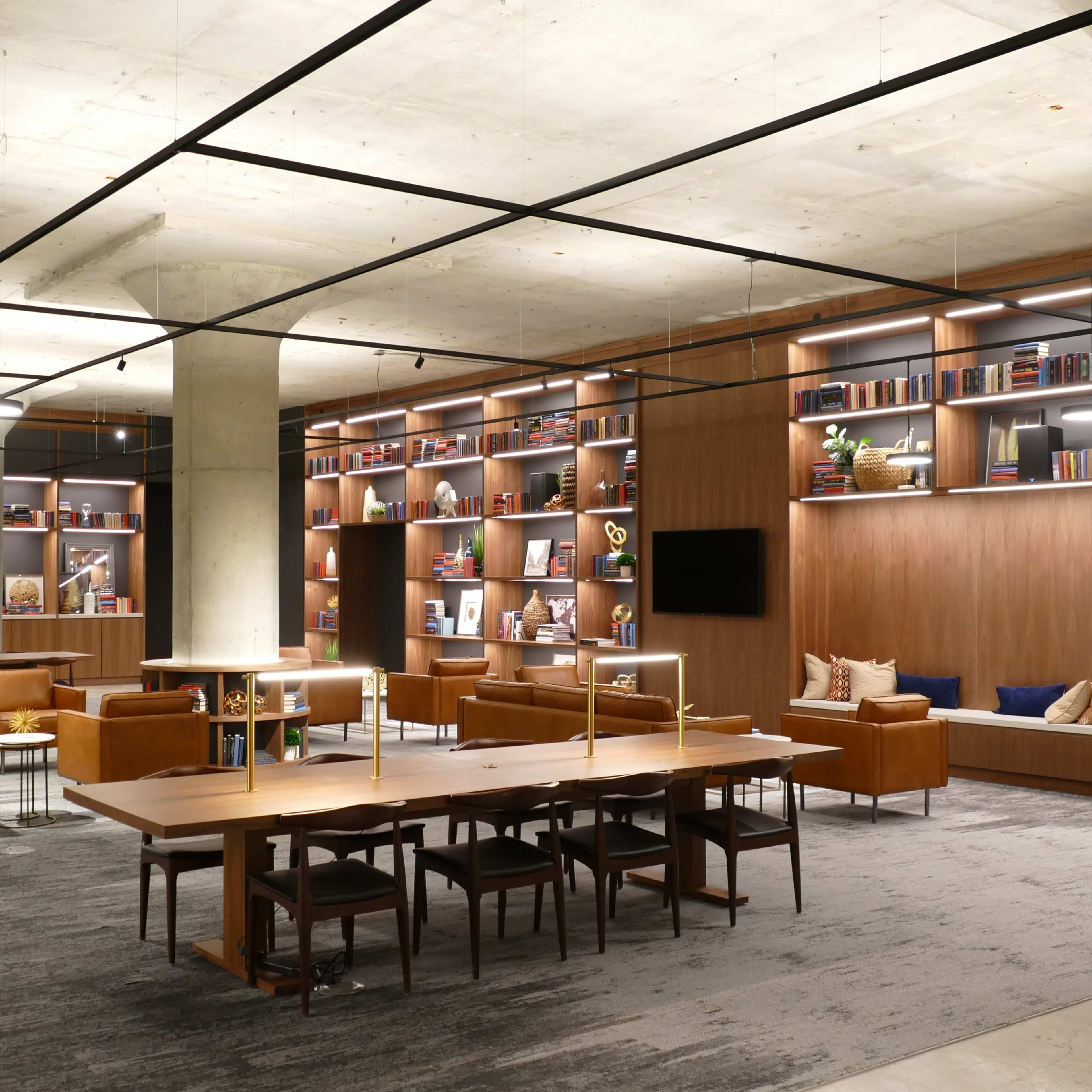 A modern, open library or lounge area with wooden shelves filled with books, seating arrangements, and a large communal table under soft lighting.