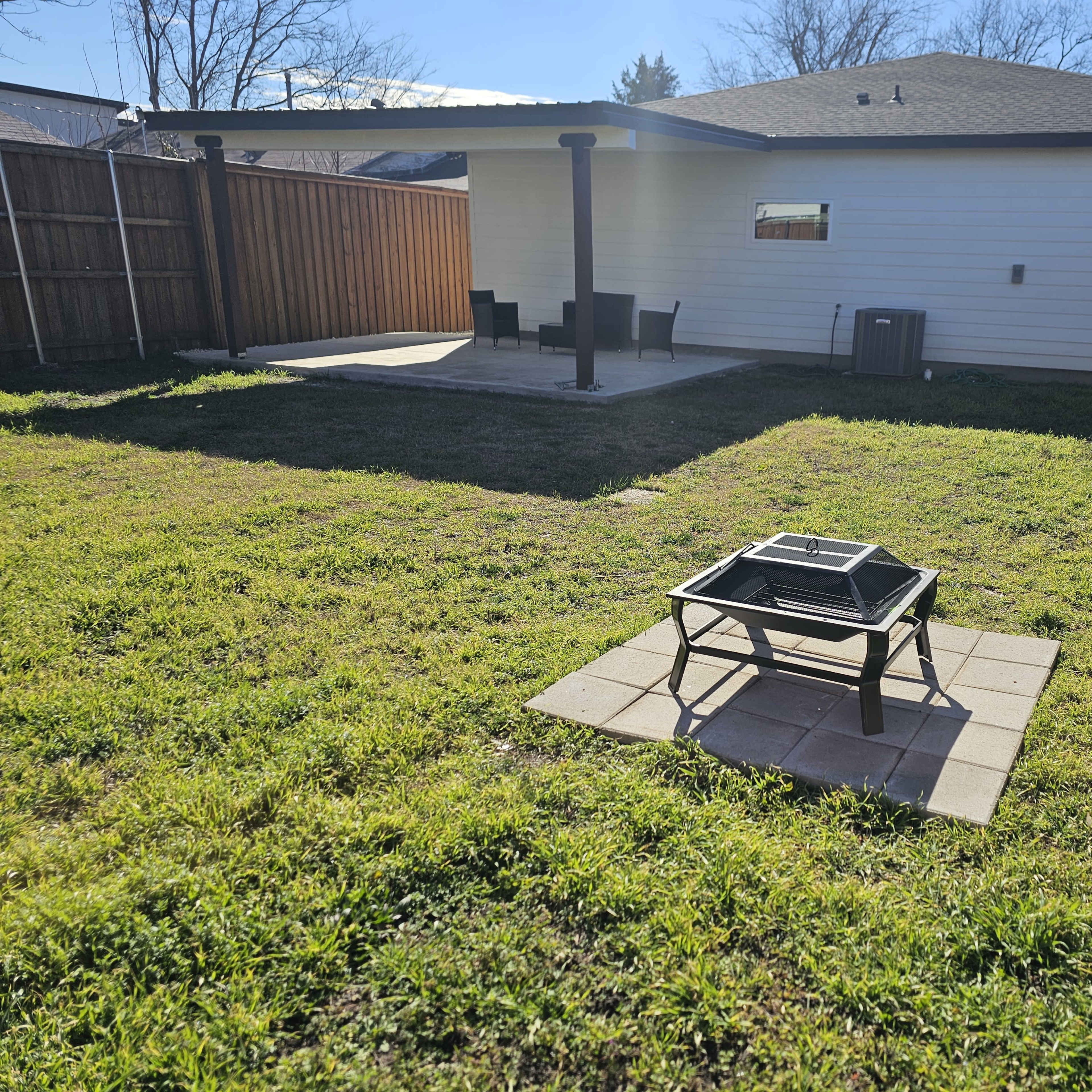 A backyard with a fire pit on a paved area, surrounded by green grass, and a house in the background with a patio and chairs.