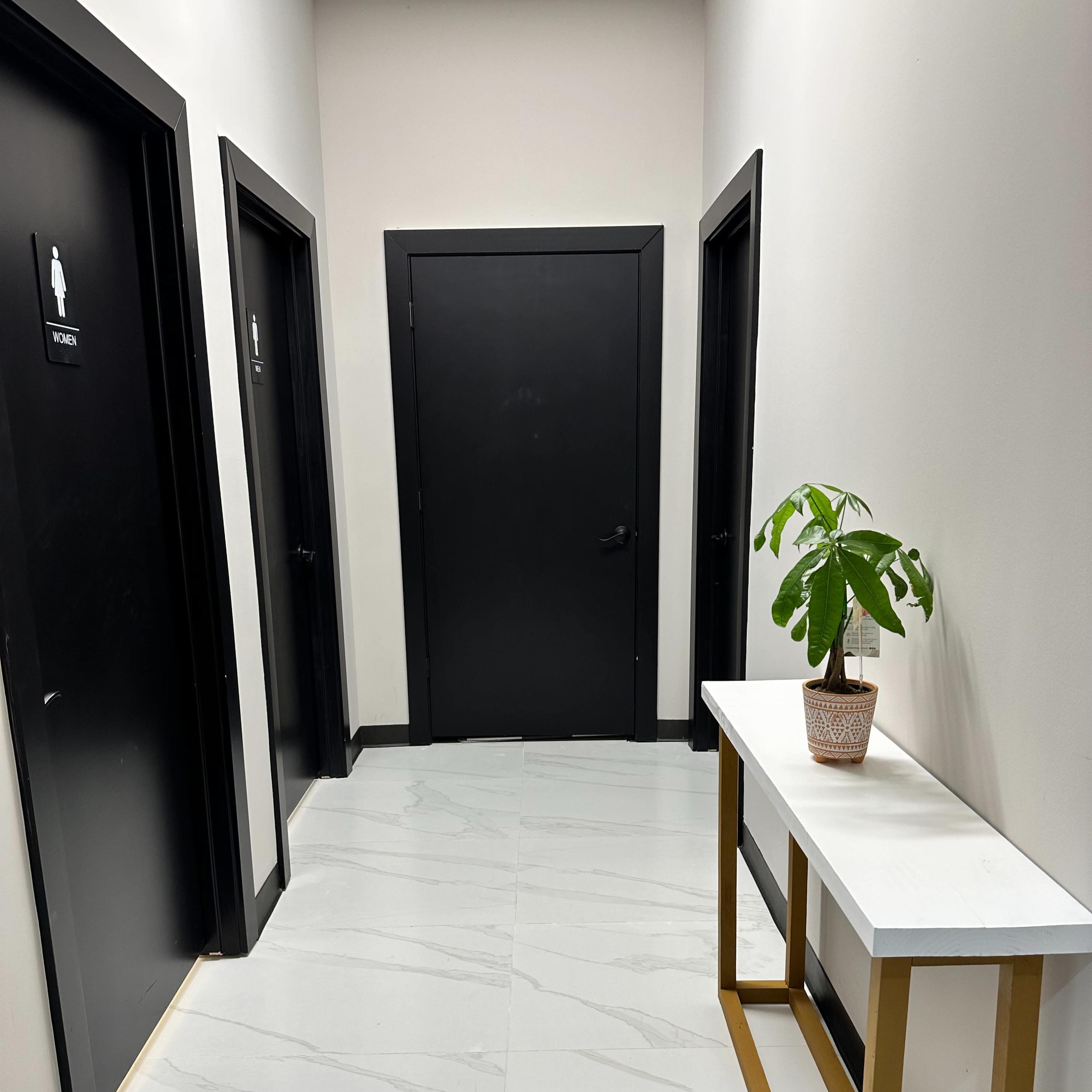 A hallway features two black restroom doors, a black door at the end, and a small table with a potted plant against a white wall.