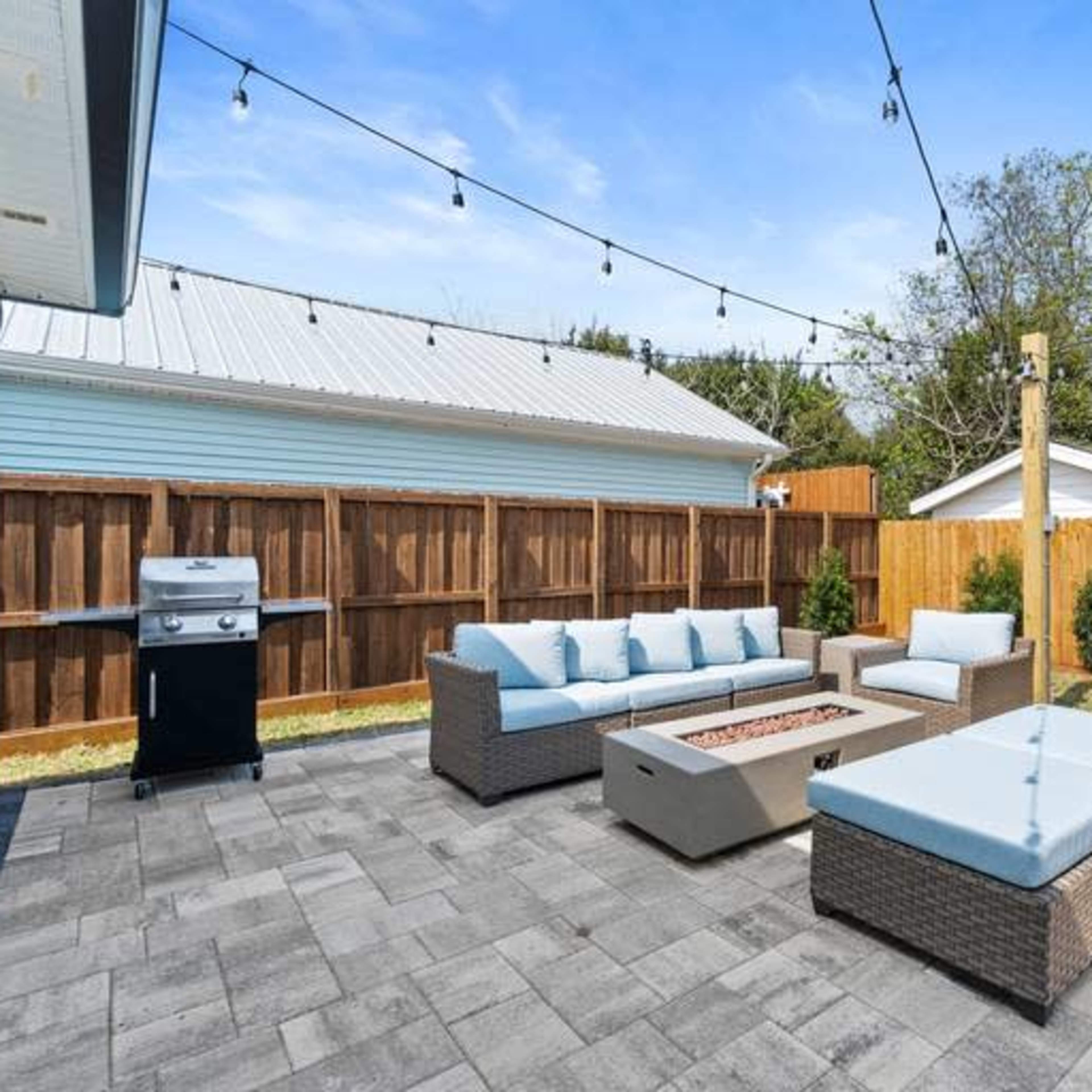A patio area features a seating arrangement with a fire pit, a grill, and string lights overhead, surrounded by a wooden fence and greenery.
