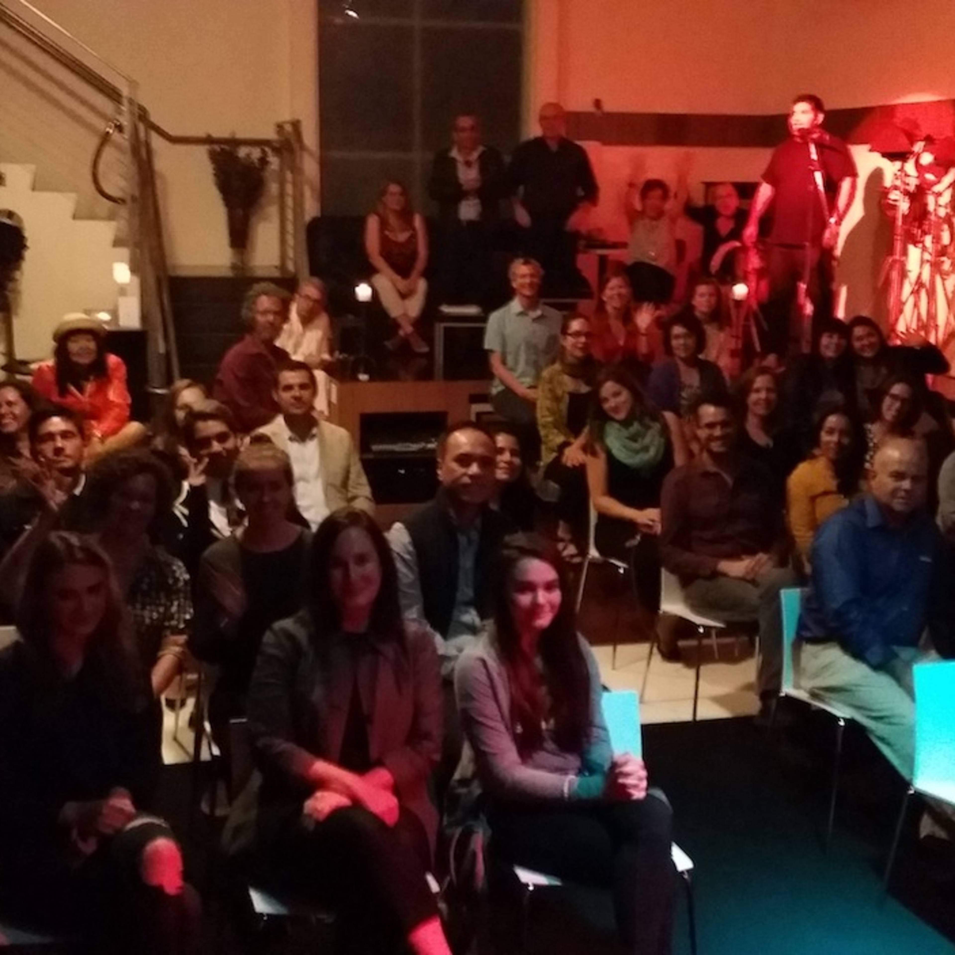 A diverse audience is seated in a dimly lit venue, with performers on a stage in the background.