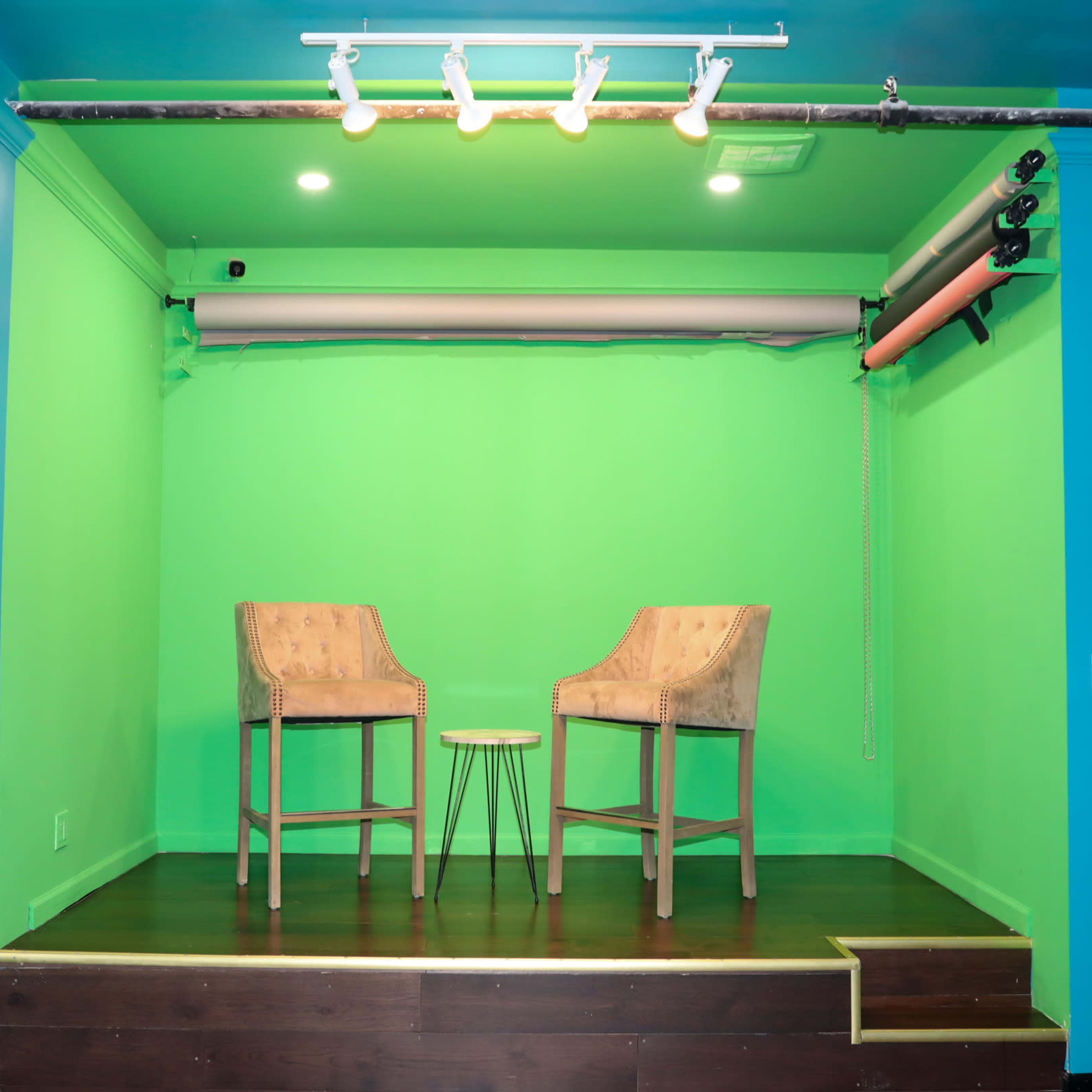 A small stage with two chairs and a table is set against a bright green backdrop.