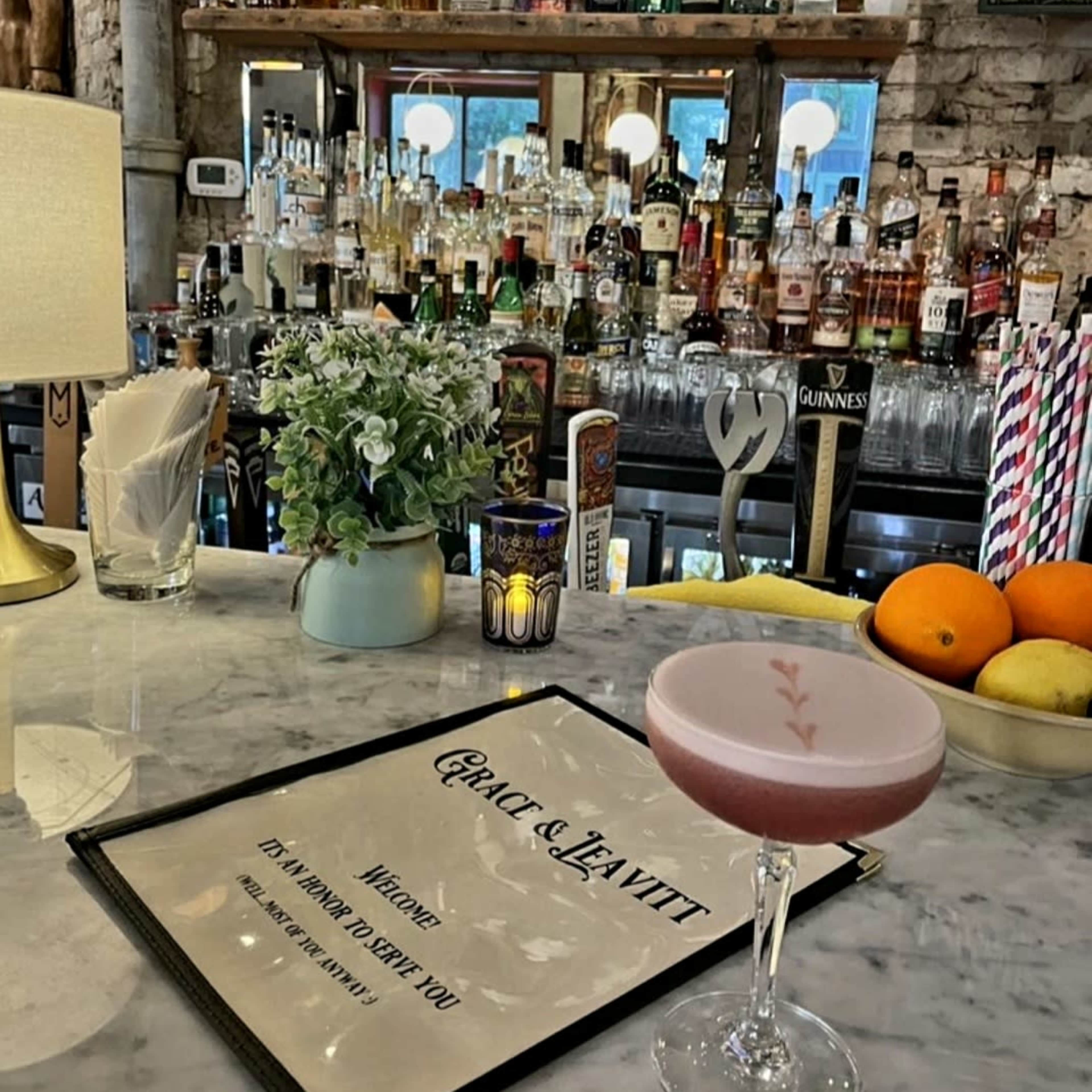 A pink cocktail sits on a marble bar counter with a menu in front of it, while a variety of liquor bottles are visible on shelves in the background.
