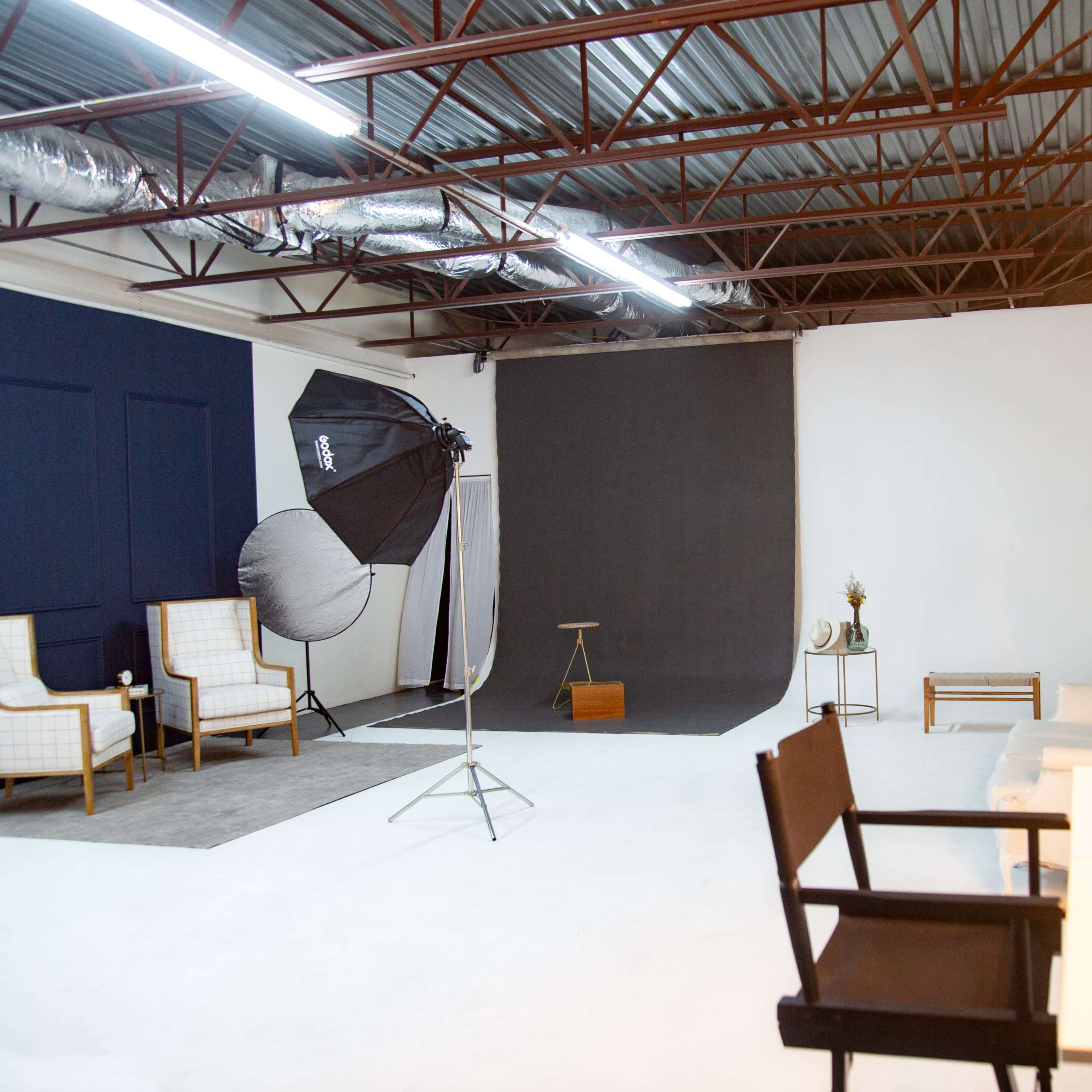 A spacious photography studio with two chairs, a large softbox light, a dark backdrop, and a vanity mirror with lights.