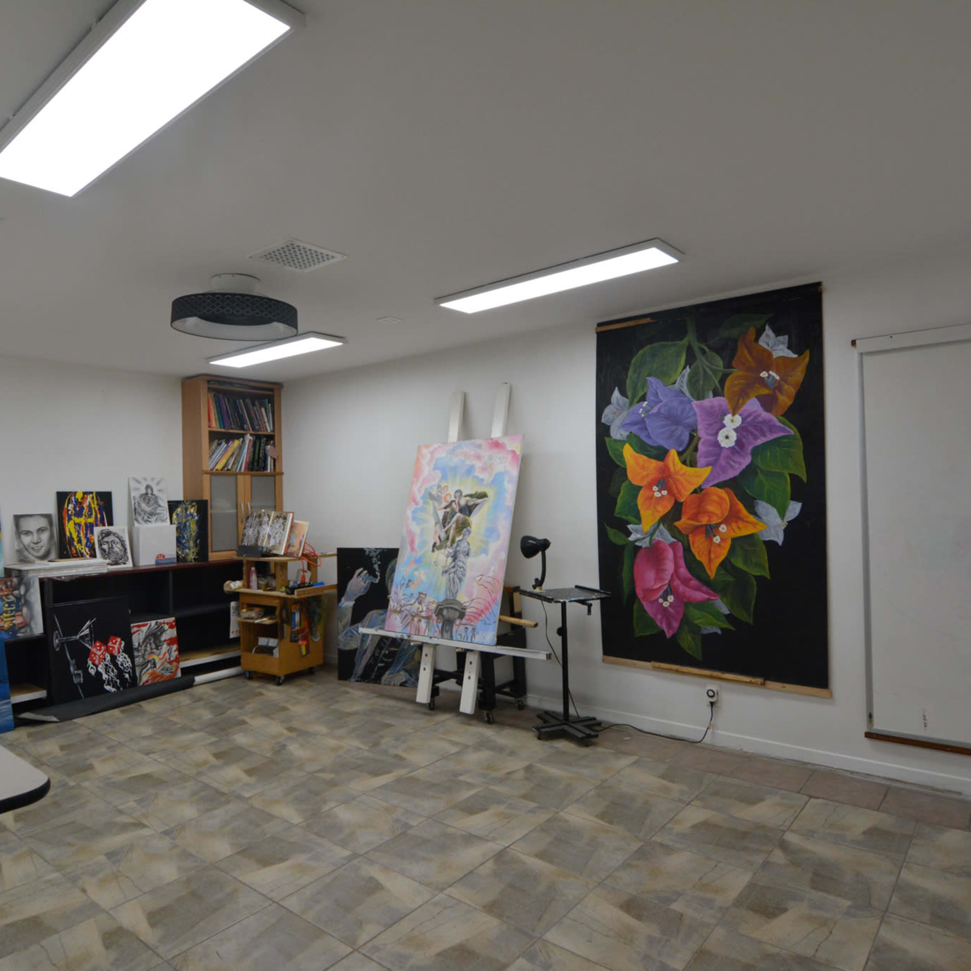 An art studio with several canvases displaying various artworks, organized shelves, and bright overhead lighting.