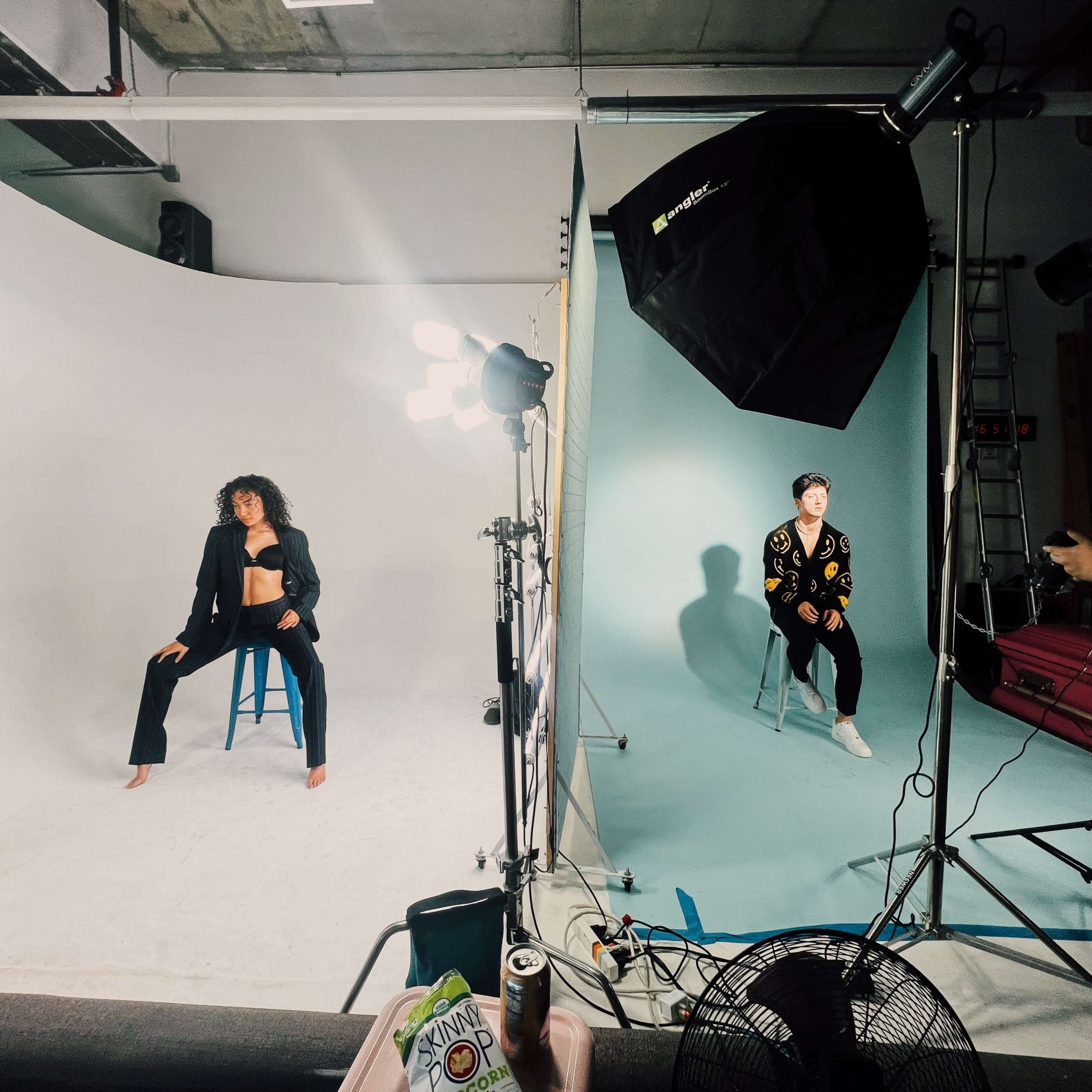 A photography studio with two models on stools, one dressed in a black outfit and the other in a colorful sweater, while a photographer adjusts lighting equipment.