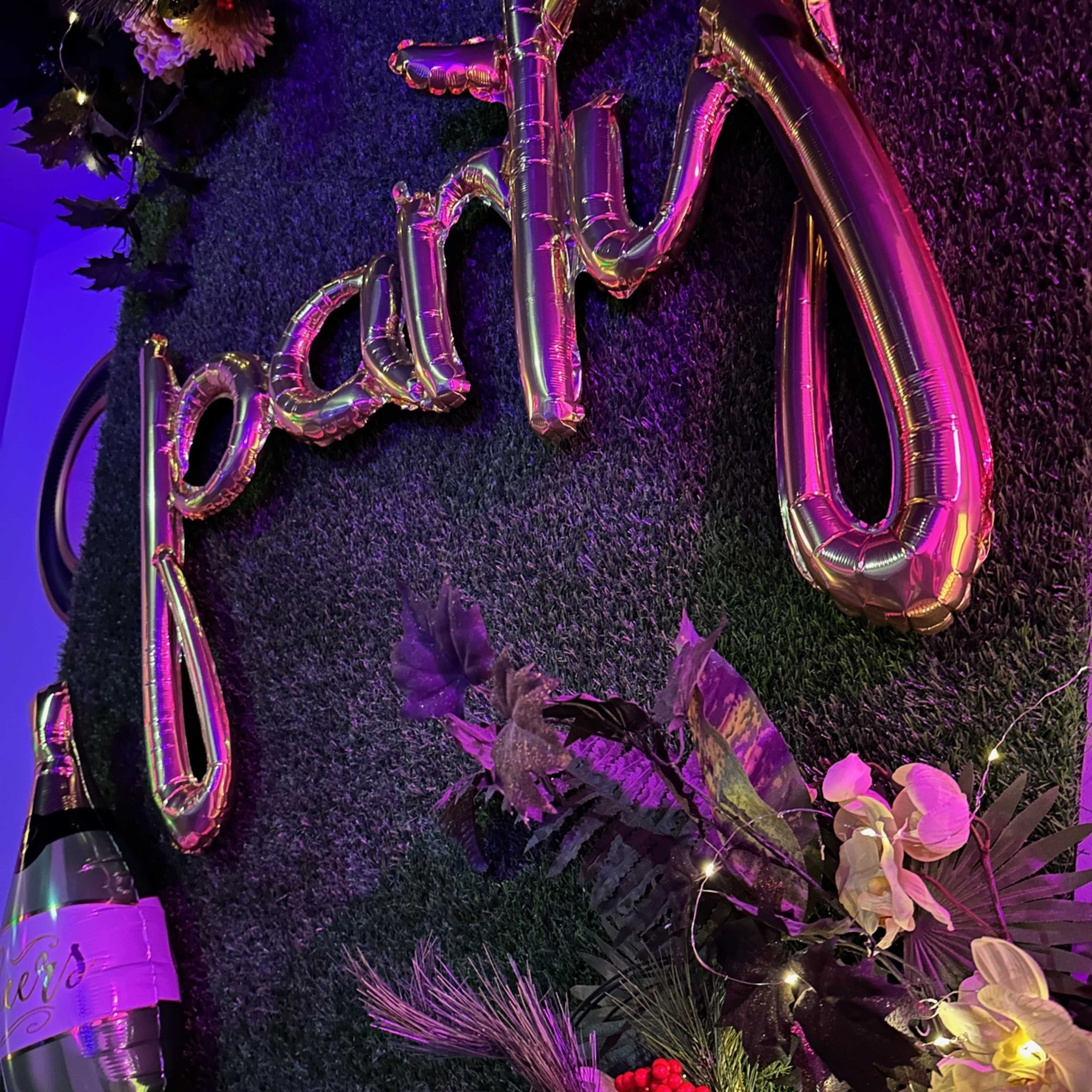 Metallic balloons spelling out "party," a bottle of champagne, and decorative flowers on a green grass-like background.