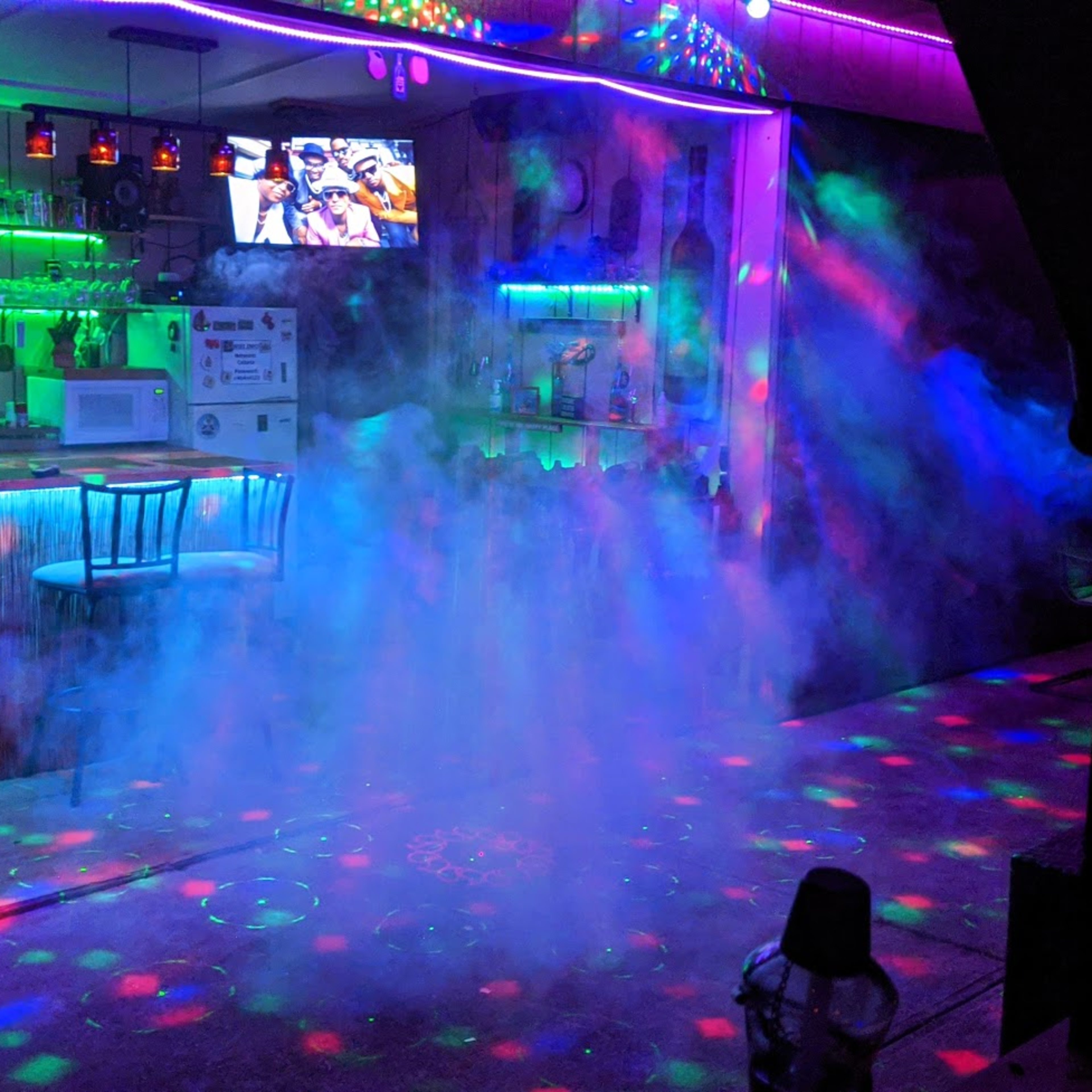 A bar area is illuminated by colorful lights and surrounded by a misty fog, creating a vibrant party atmosphere.