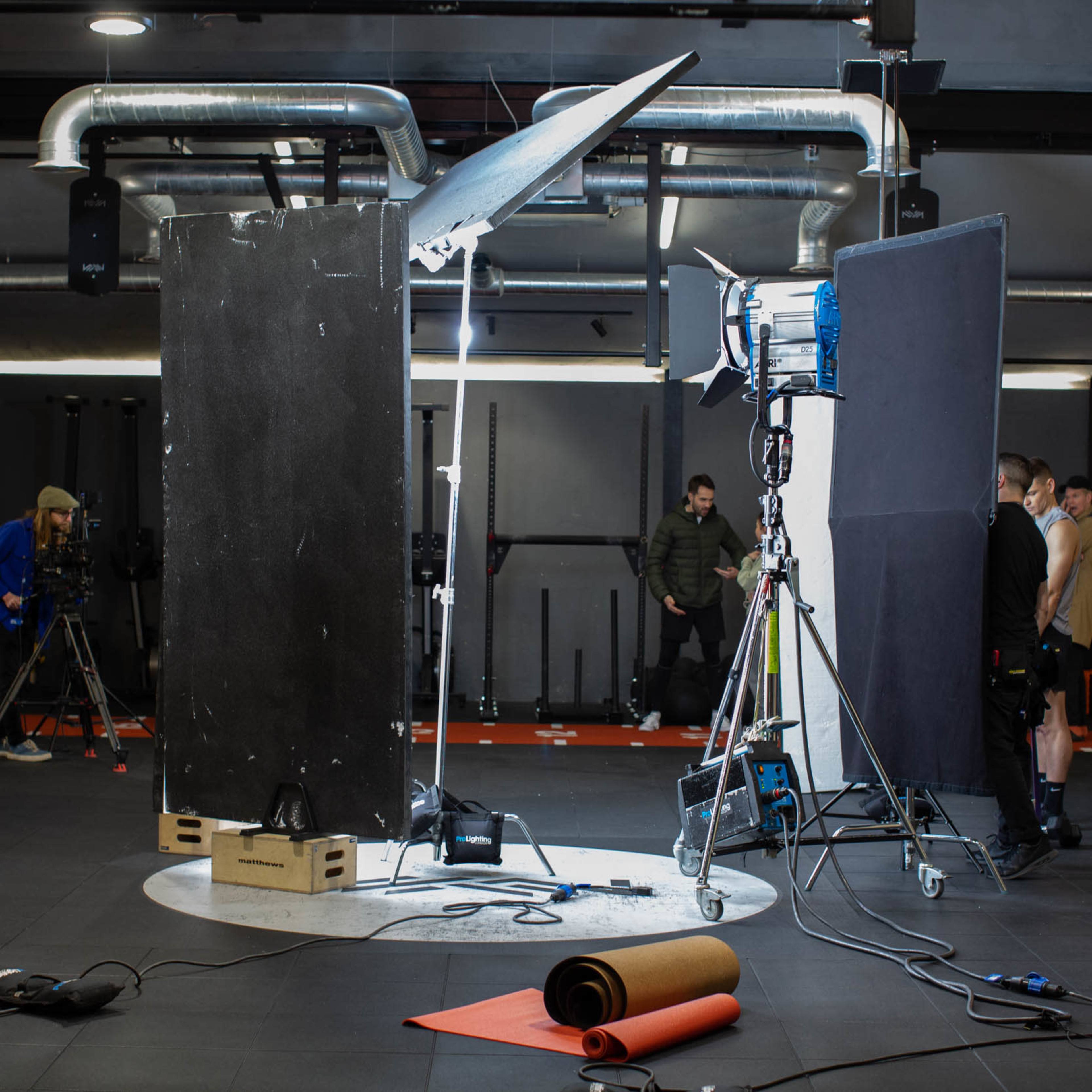 A photo studio setup features lighting equipment and reflectors in a spacious gym environment, with several individuals behind the scenes and exercise equipment in the background.