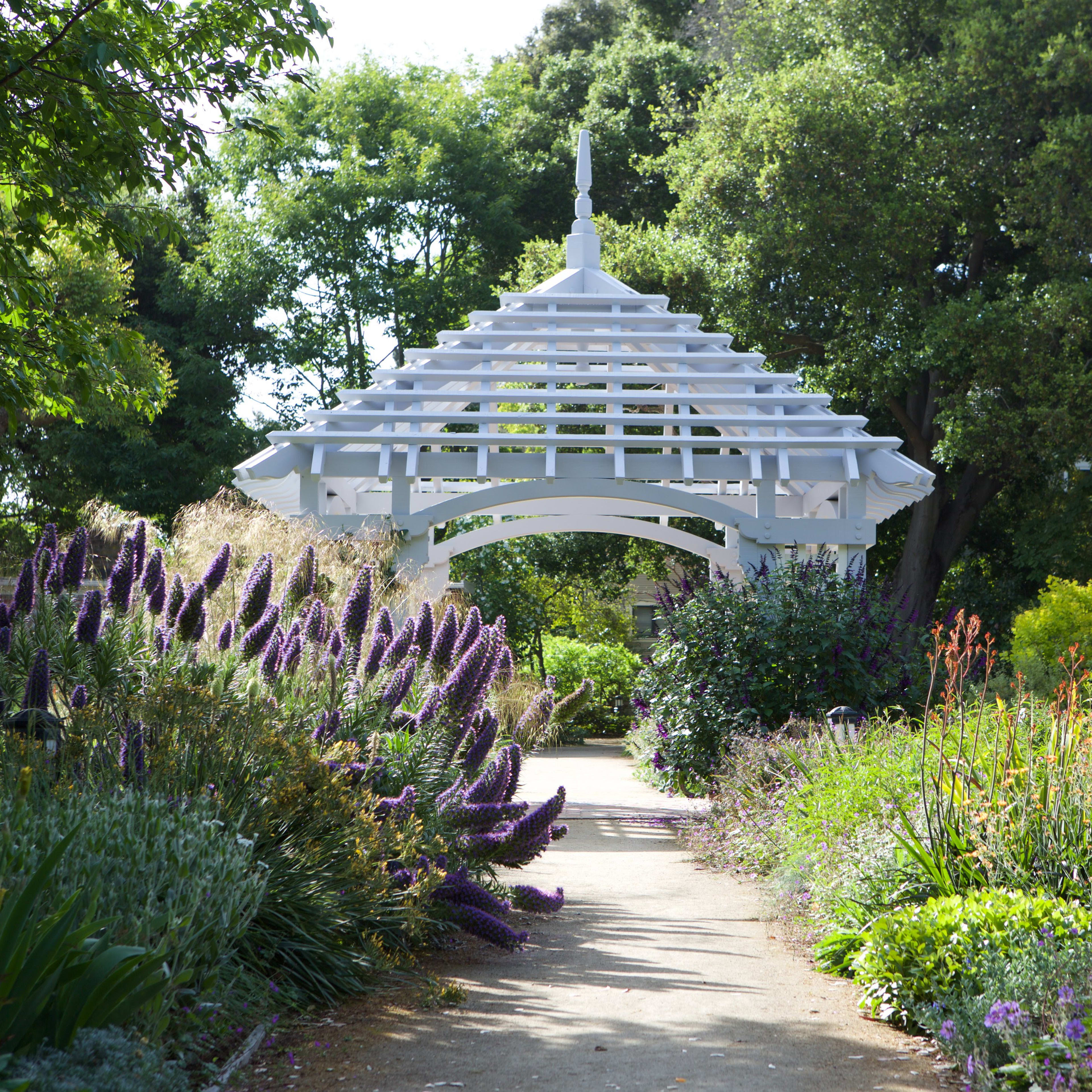 A white wooden gazebo is situated at the end of a pathway lined with colorful flowering plants and lush greenery.