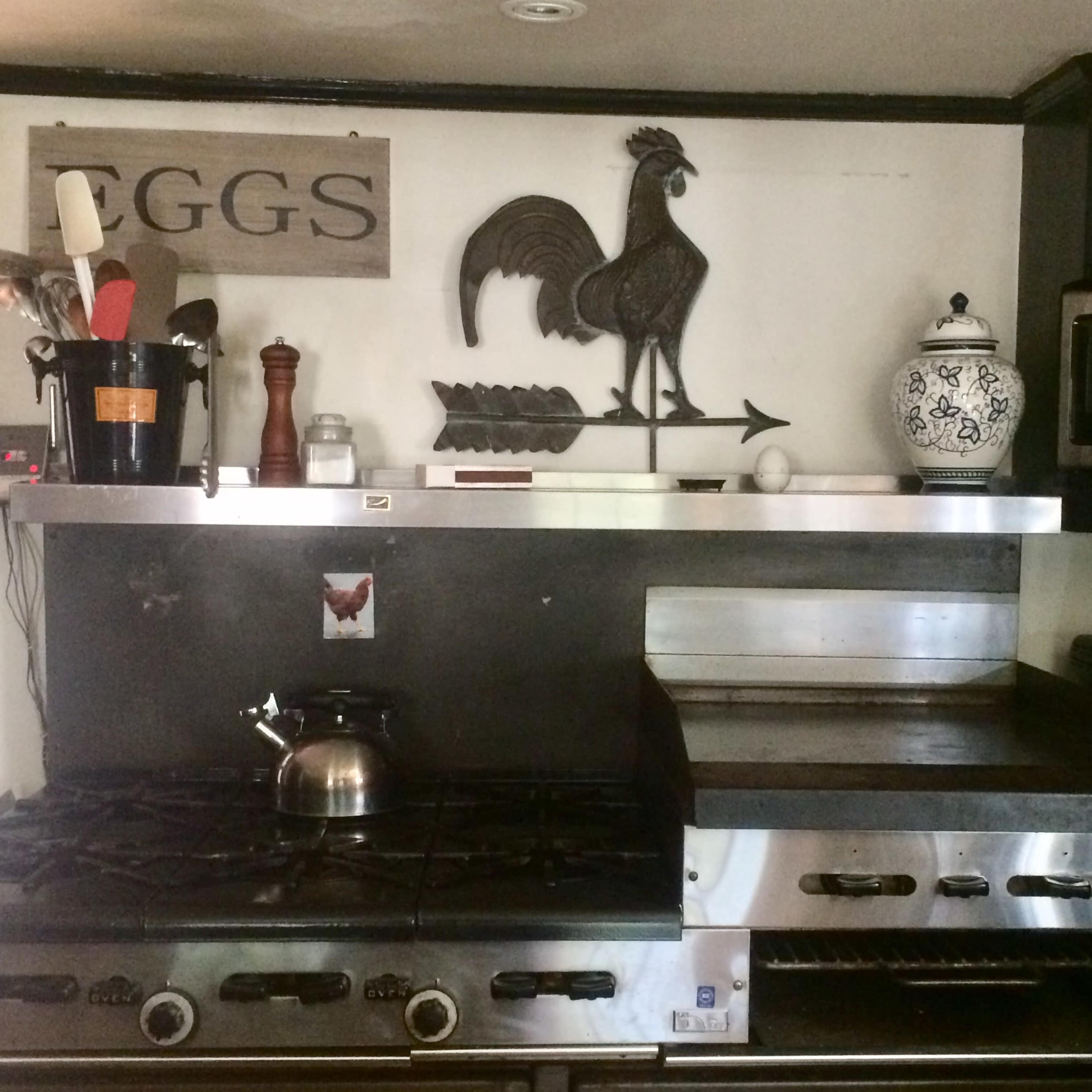 A kitchen with a gas stove, a metal griddle, cooking utensils in a container, and decorative items including a rooster weather vane and various containers on the shelves.